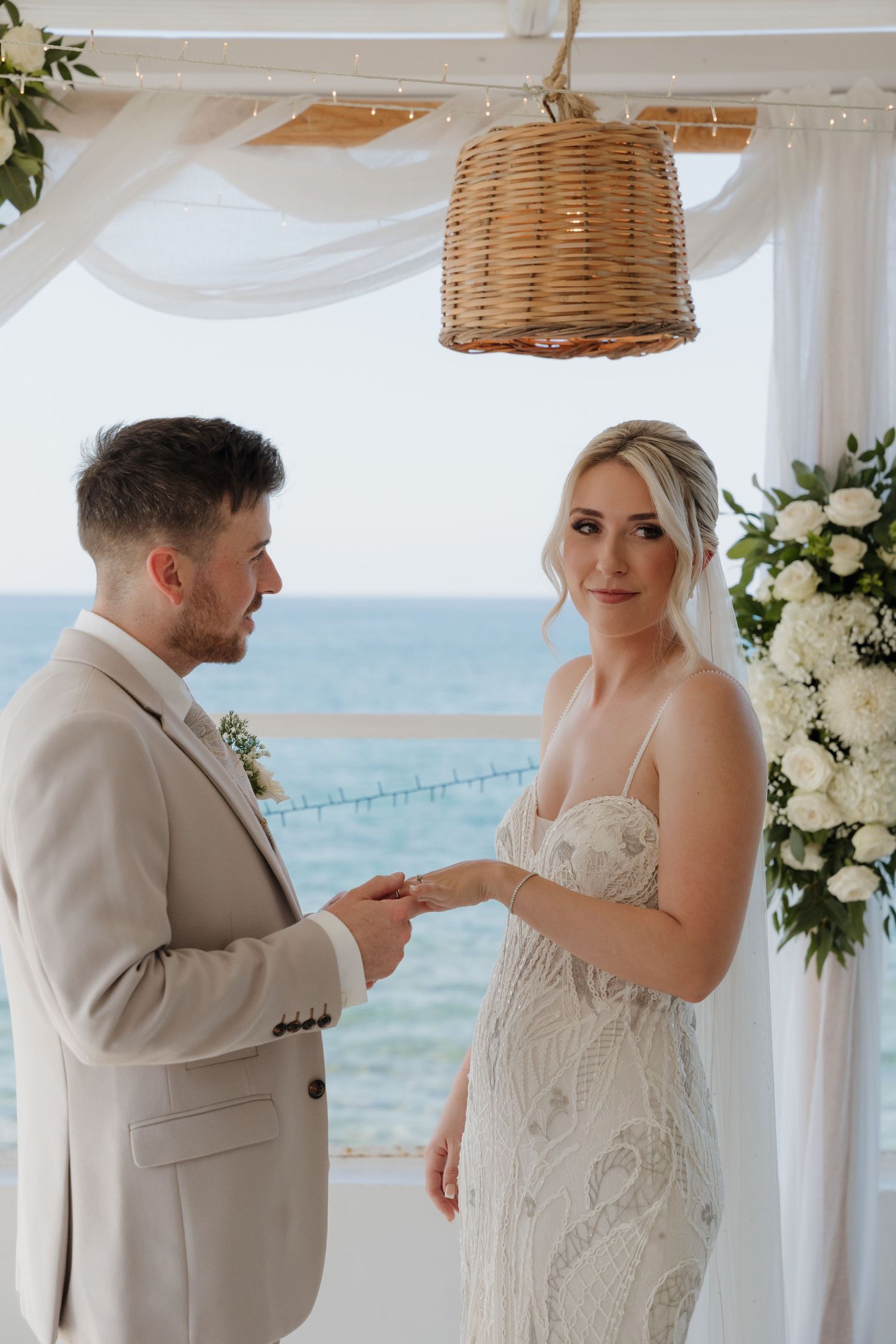 A wedding ceremony at an oceanfront venue with a woven pendant light and white floral arrangements.