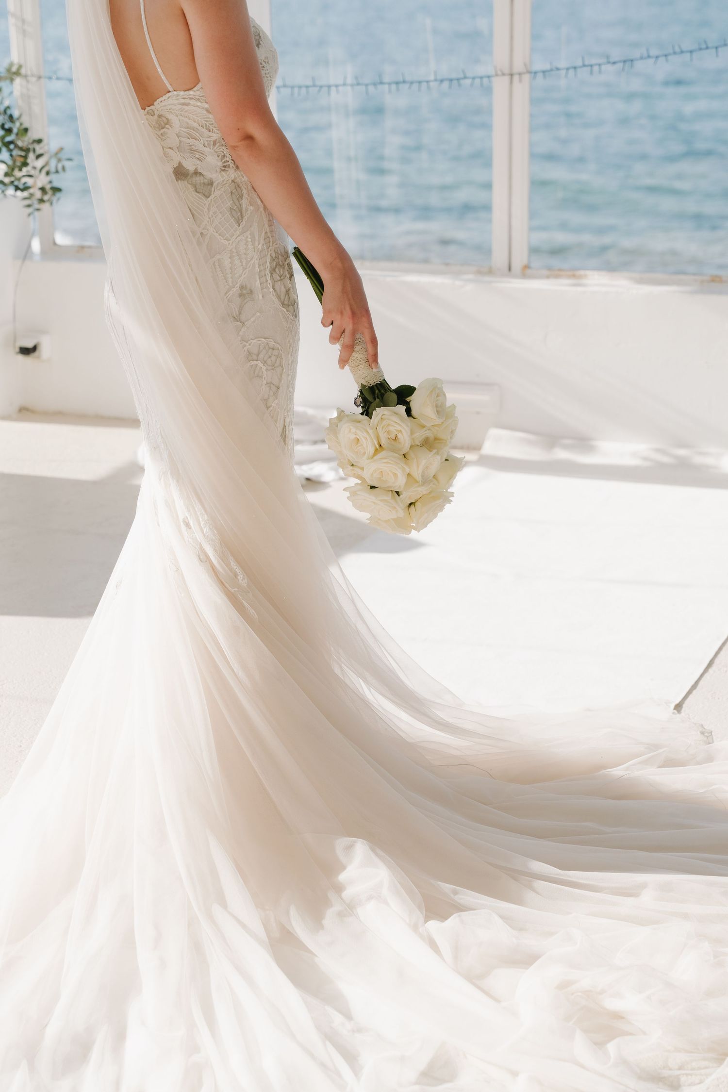 Close-up detail of a flowing white wedding dress train with delicate lace details and white bouquet.