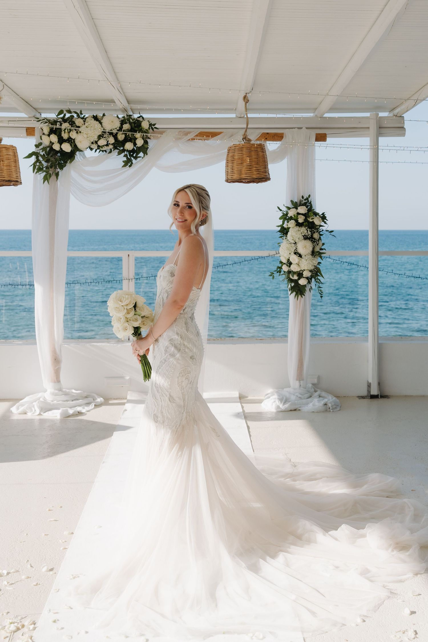 Stunning bride in a fitted lace gown poses on an oceanfront terrace with floral decorations.