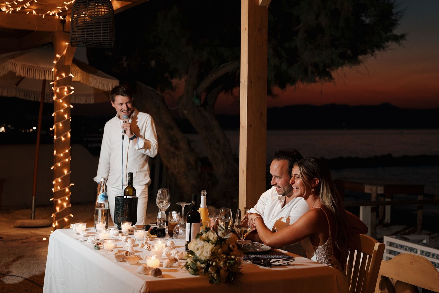 Evening wedding reception with string lights as guest gives speech at decorated oceanside table.