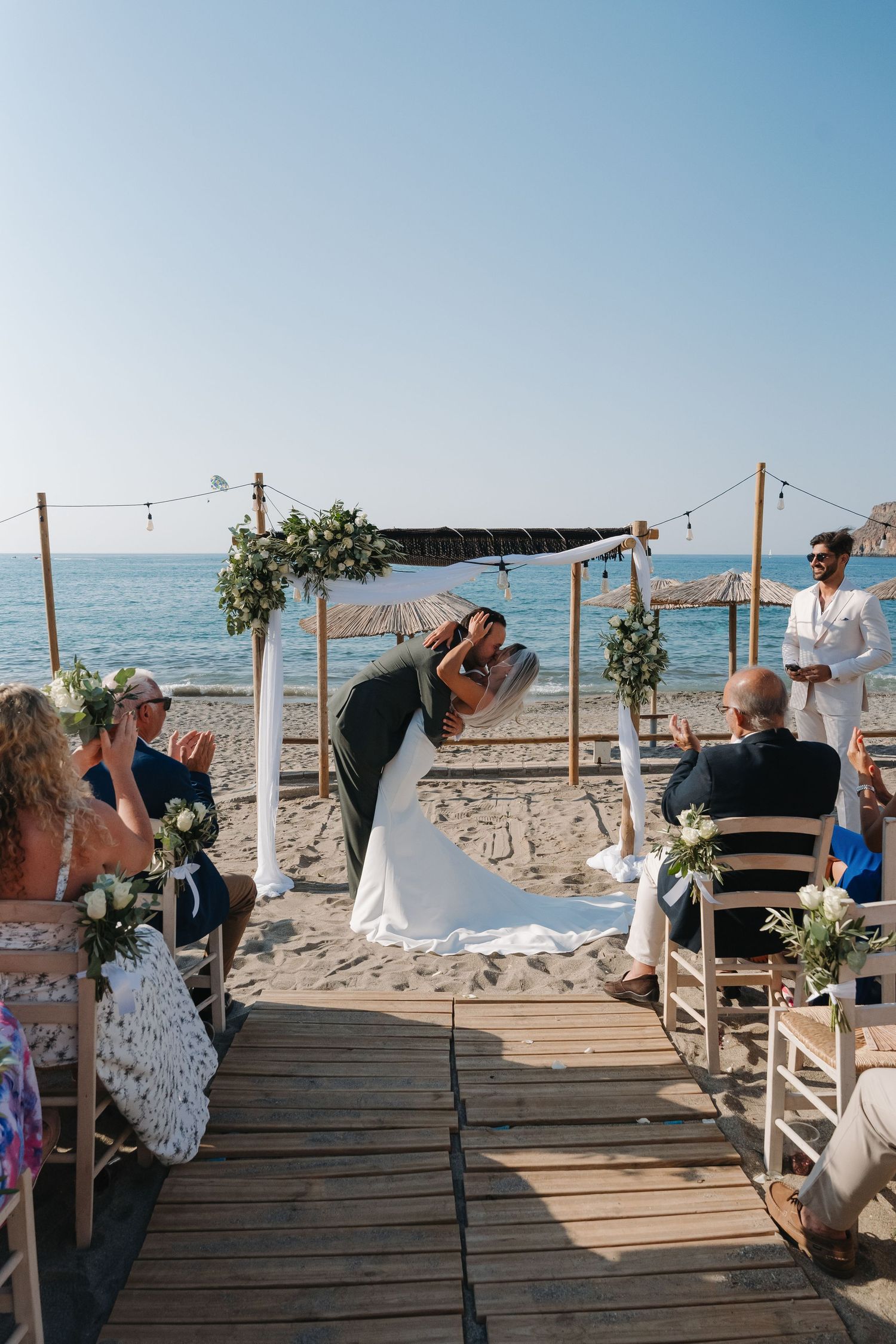 Guests seated at a rustic beachfront wedding ceremony with wooden boardwalk and ocean views.