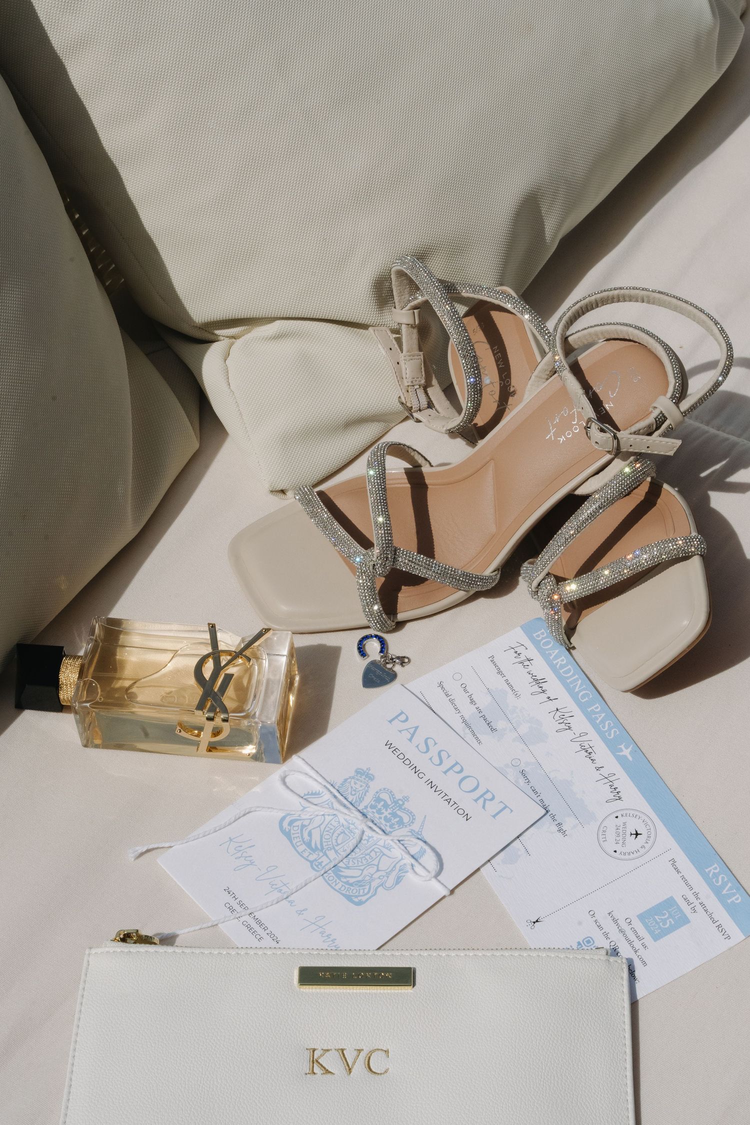 Wedding accessories including YSL perfume, silver sandals, passport and white clutch on neutral background.