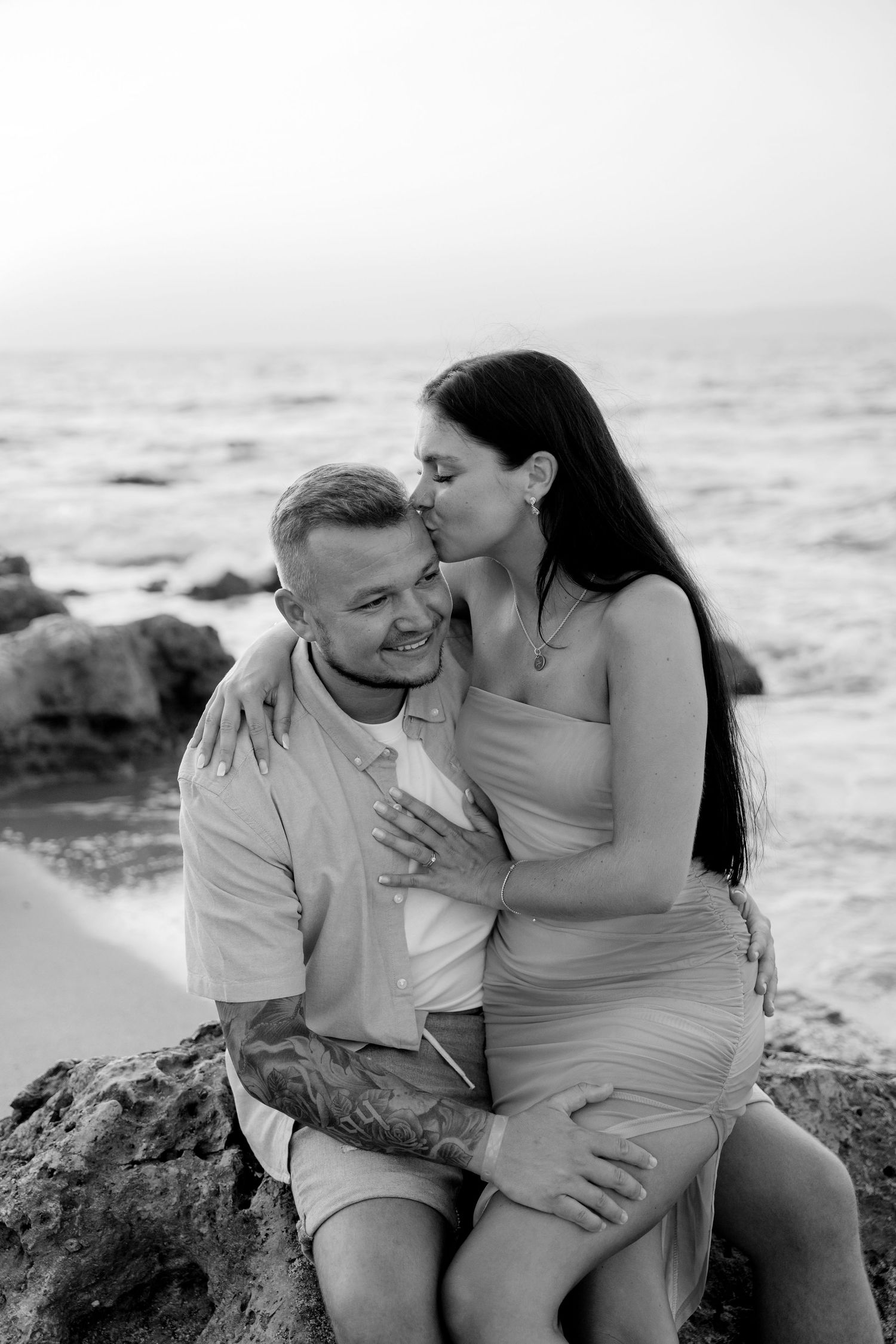 A black and white photo of a couple sharing a tender moment on coastal rocks.