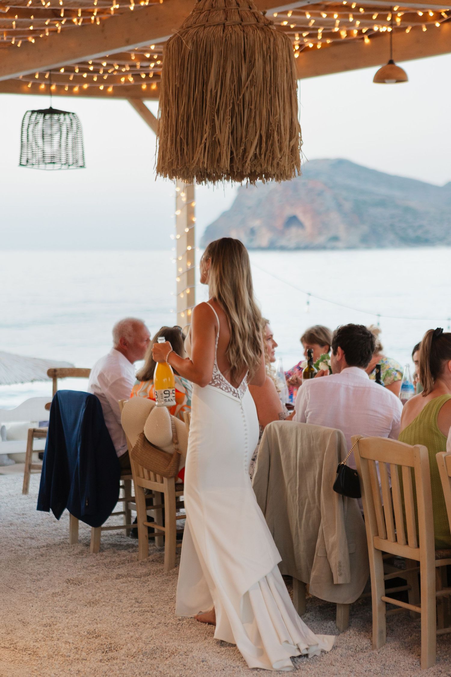 Bride in fitted white dress stands near patio dining area overlooking coastal mountain views.