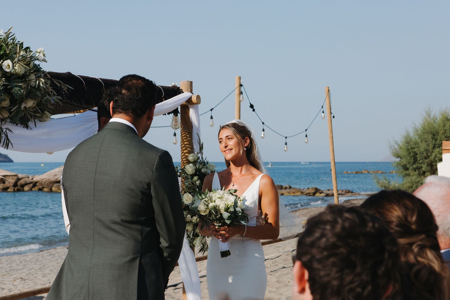 Bride and groom exchange vows during intimate waterfront ceremony with string lights overhead.