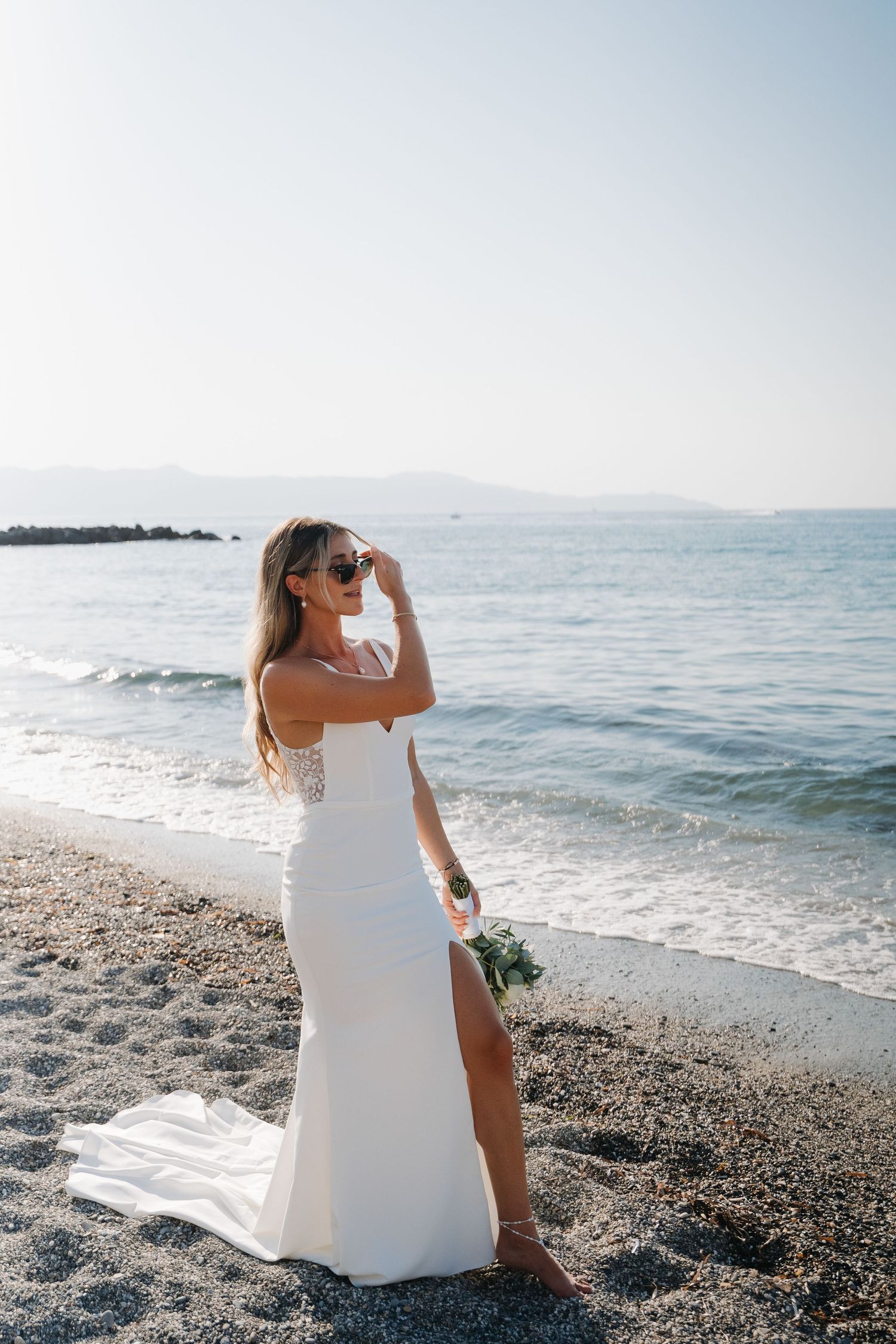 Bride in sleek white dress with thigh slit poses on beach with scenic ocean view.