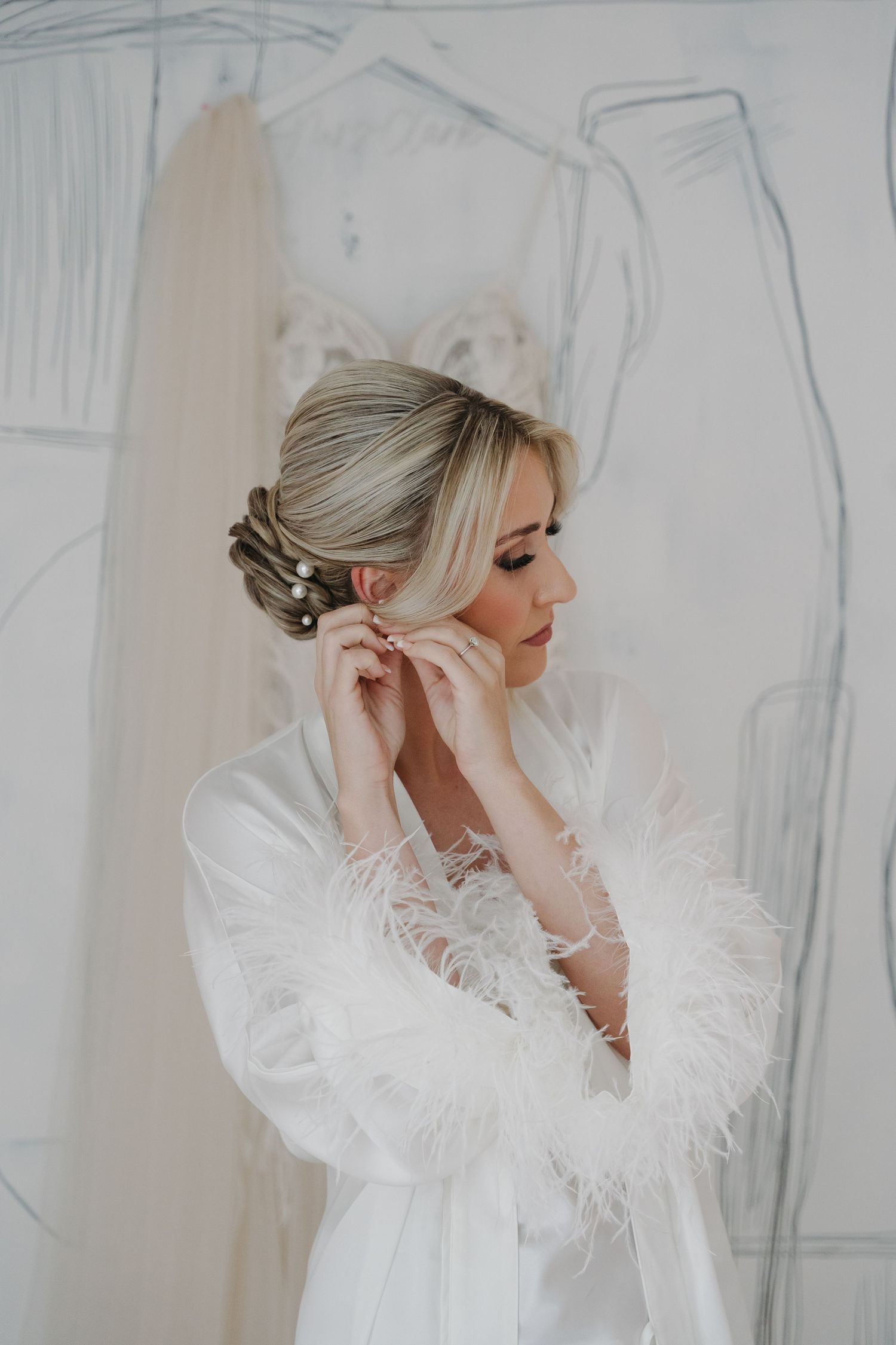 A person in a white feathered robe putting on earrings during wedding preparations with elegant updo hairstyle.