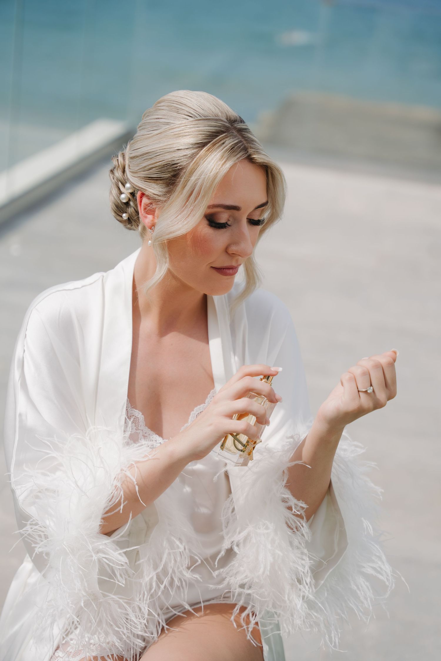 Elegant bride in feathered robe applying perfume with ocean view.