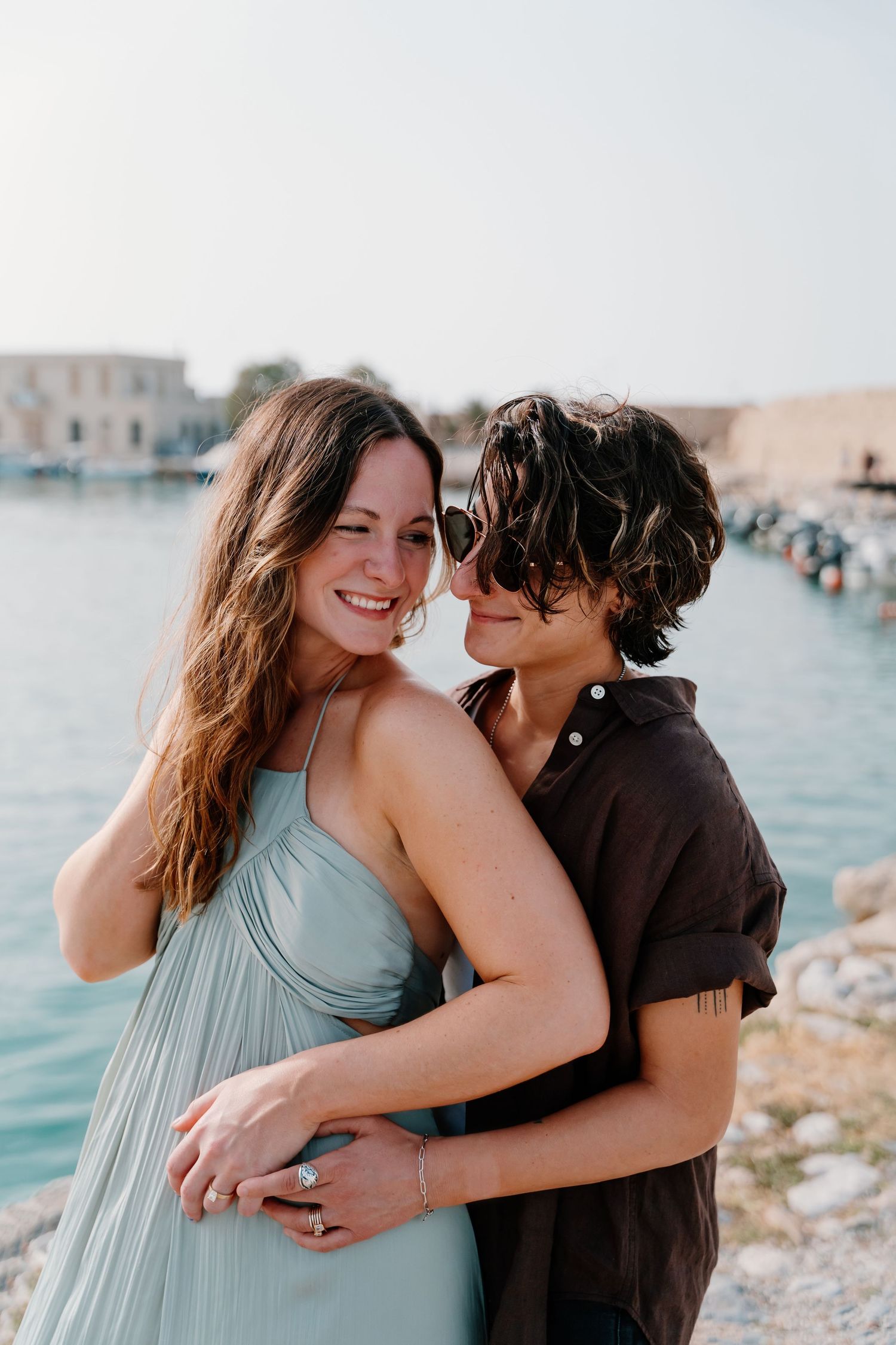 A romantic couple embraces by turquoise waters along a Mediterranean coastline in Rethymno, Crete on a sunny afternoon.