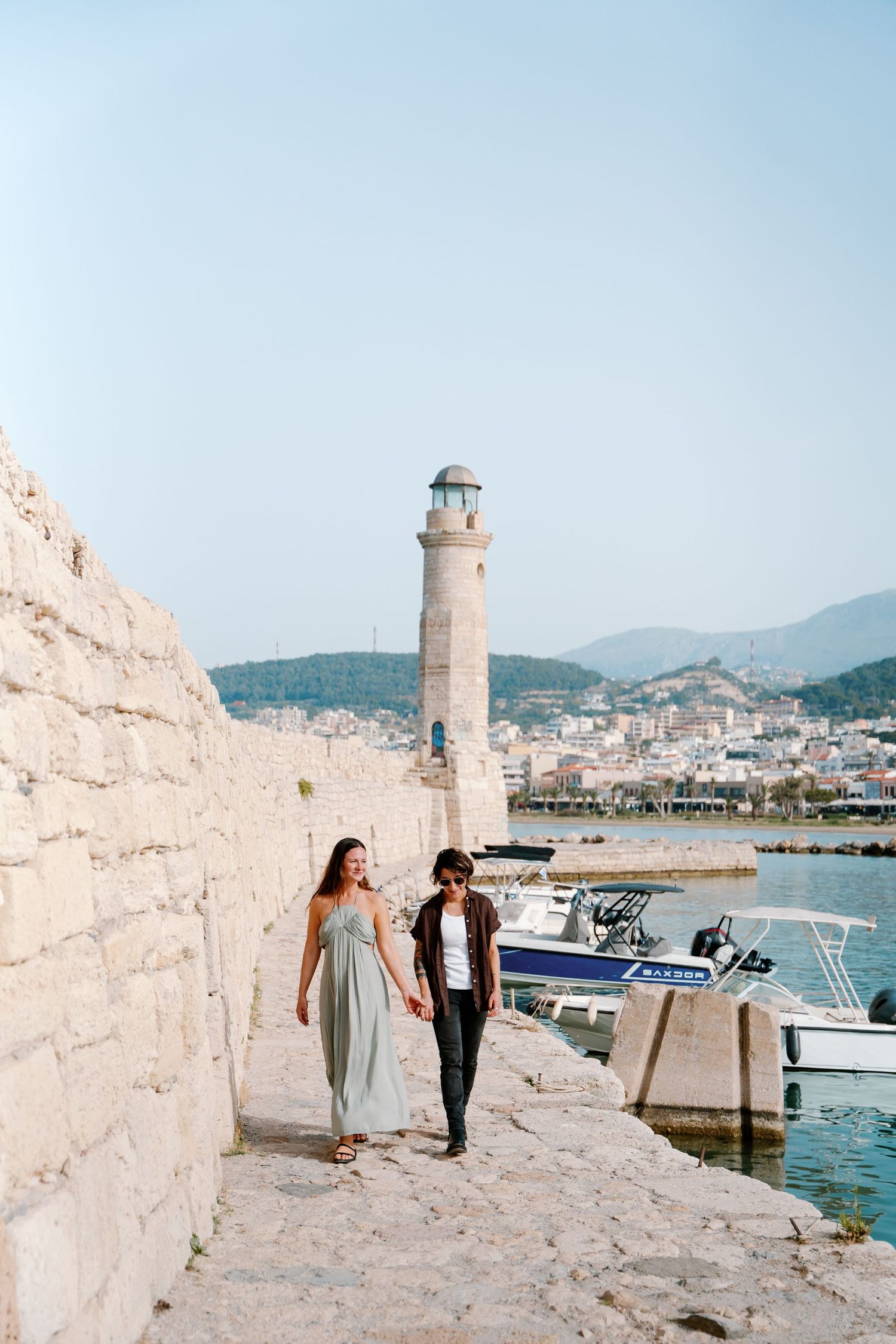 A couple strolls along a scenic harbor path in Rethymno, Crete with a white lighthouse and boats in the Mediterranean coastal backdrop.