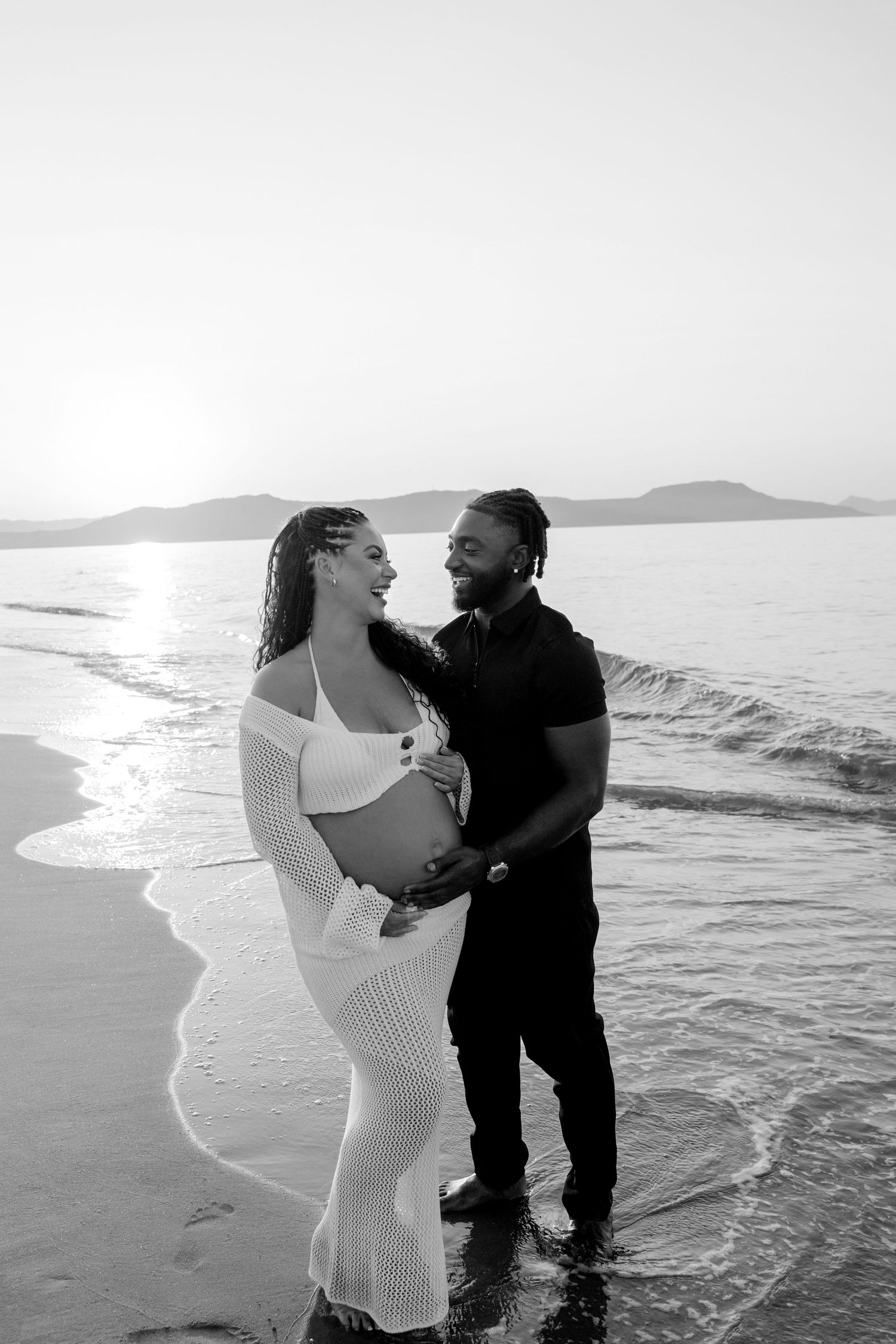 Couple embraces during beachside maternity photoshoot with waves rolling in behind them.
