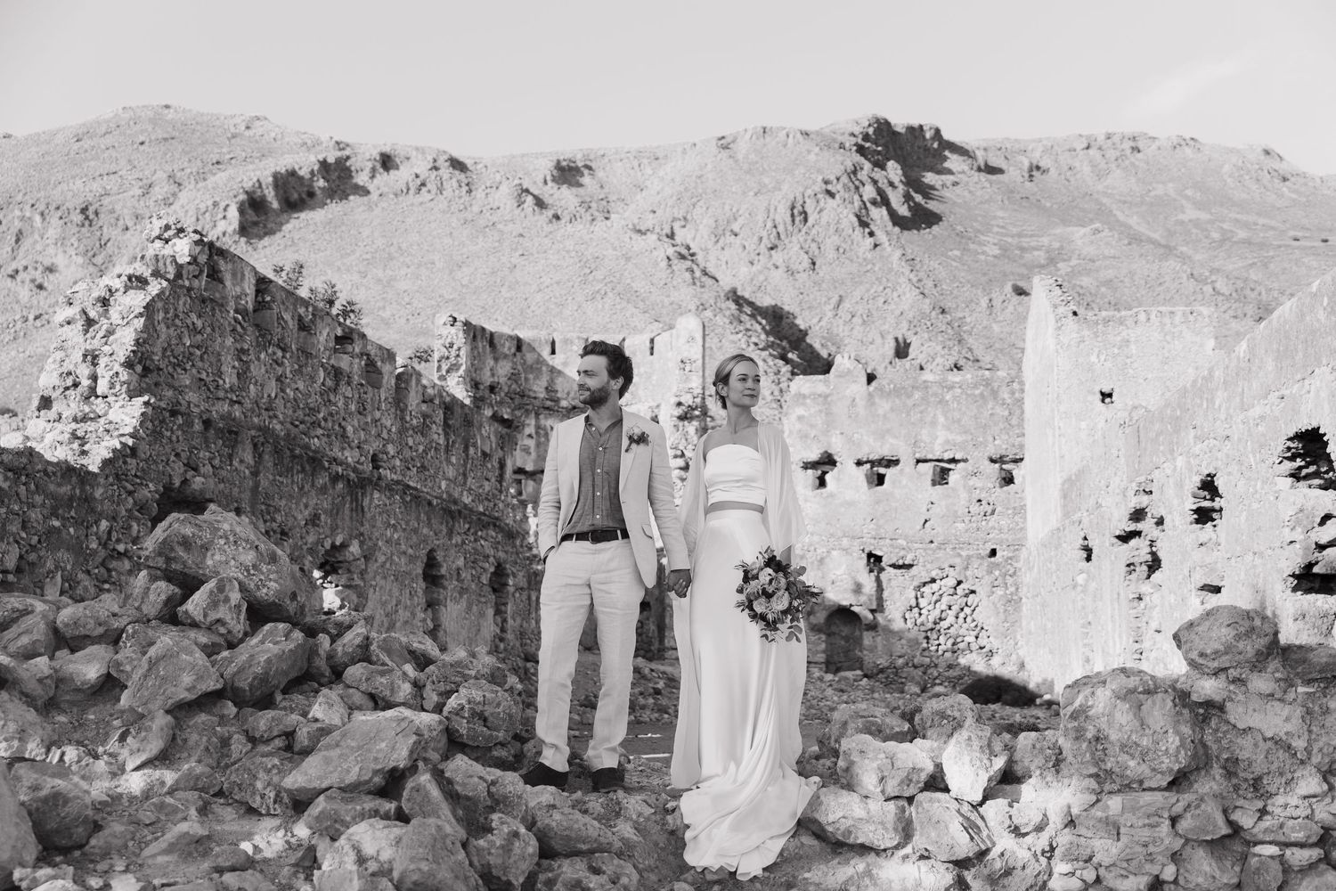 A couple stands among ancient stone ruins in a mountainous landscape in Loutro, Crete captured in black and white photography.
