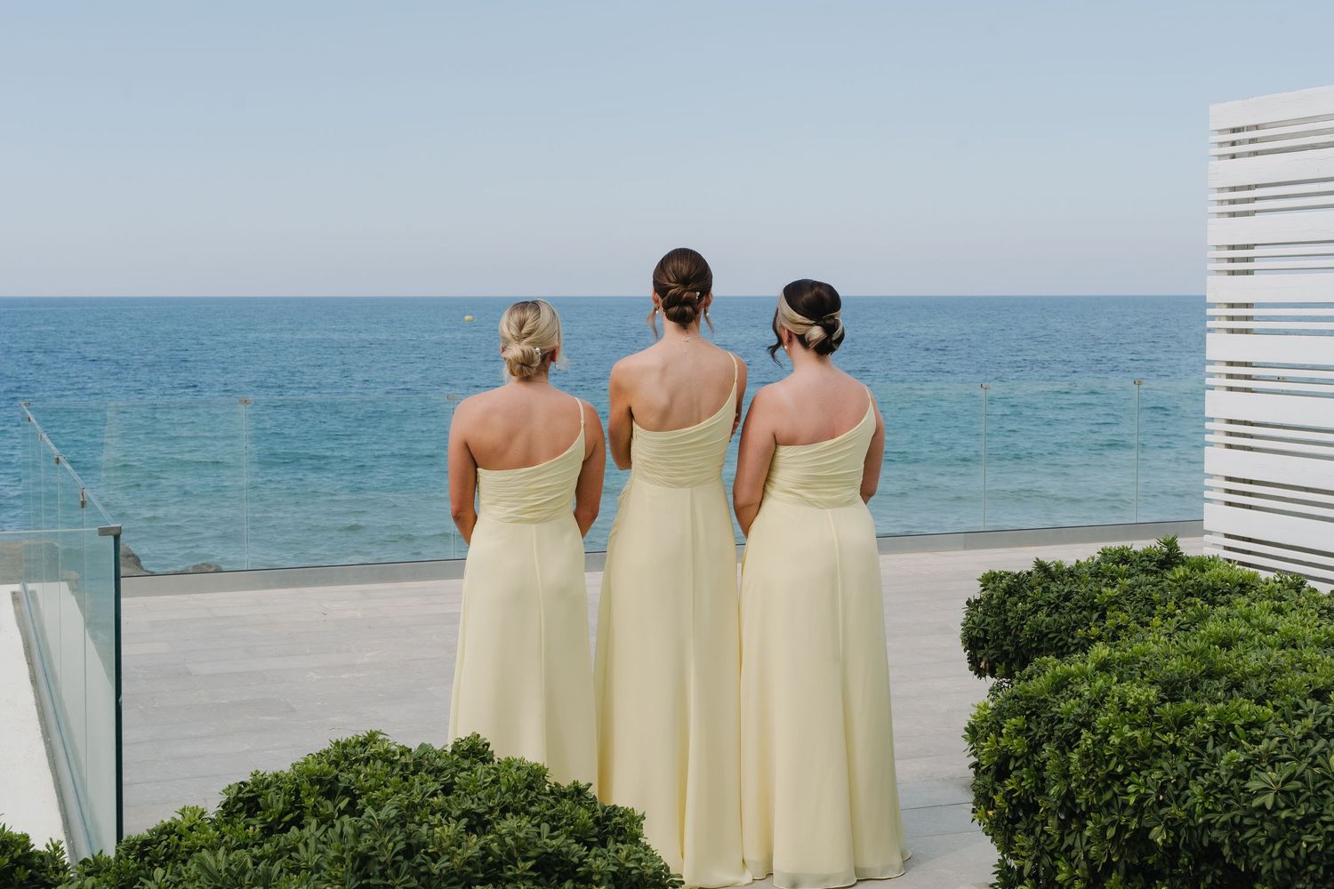 Three bridesmaids in pale yellow dresses overlooking ocean view from seaside wedding venue.