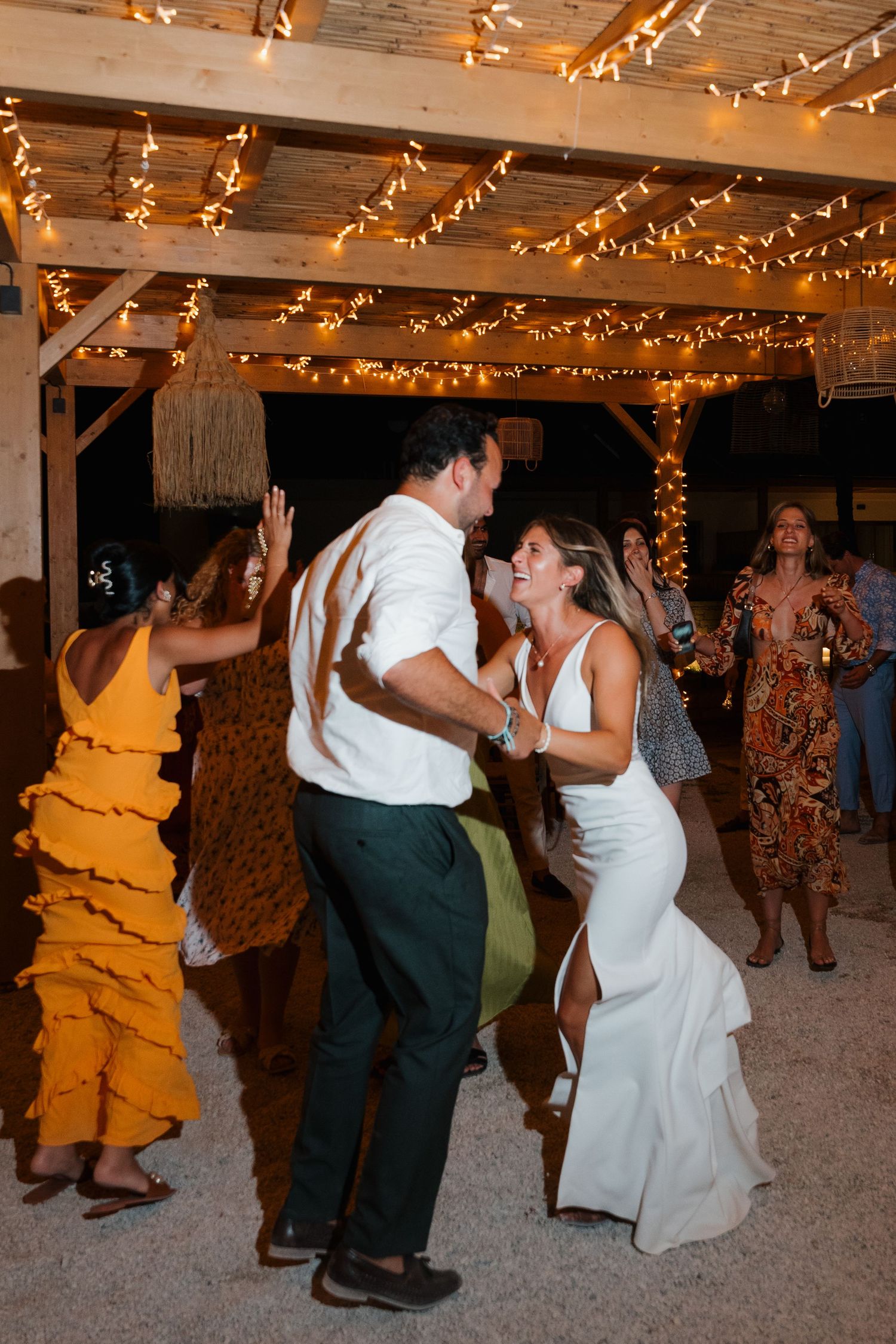 Wedding guests dance and celebrate under wooden beams decorated with warm string lights at an outdoor reception.