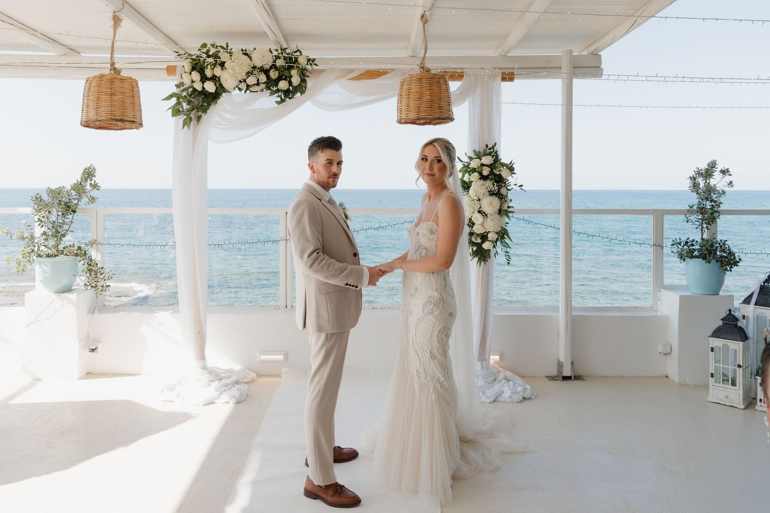 Bride and groom standing together on white oceanfront terrace decorated with flowers.
