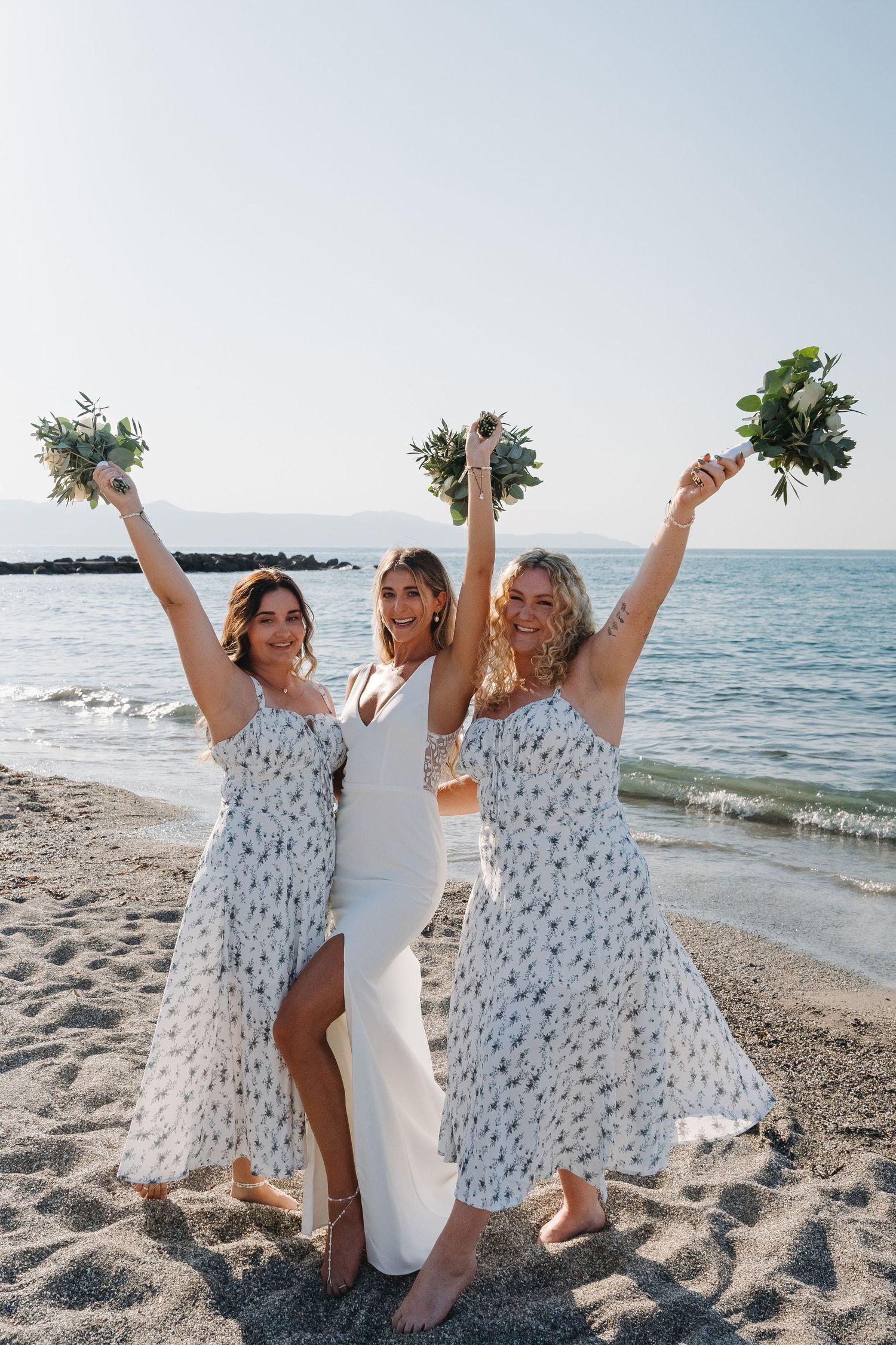 Three bridesmaids in matching floral dresses celebrate joyfully on a sunny beach with raised bouquets.
