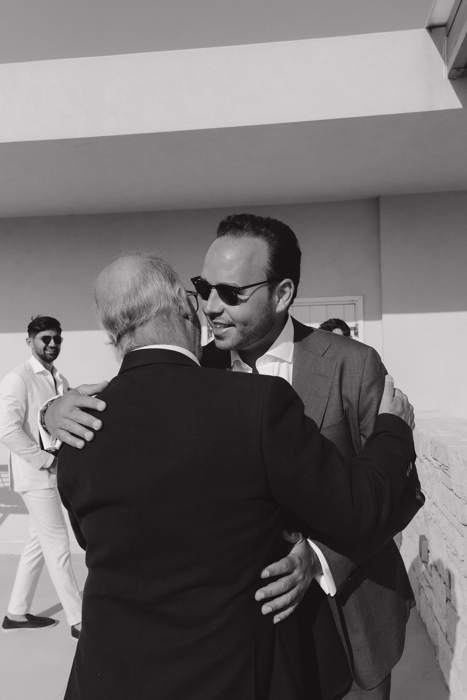 Two men in formal attire embrace at an outdoor event in black and white photography.