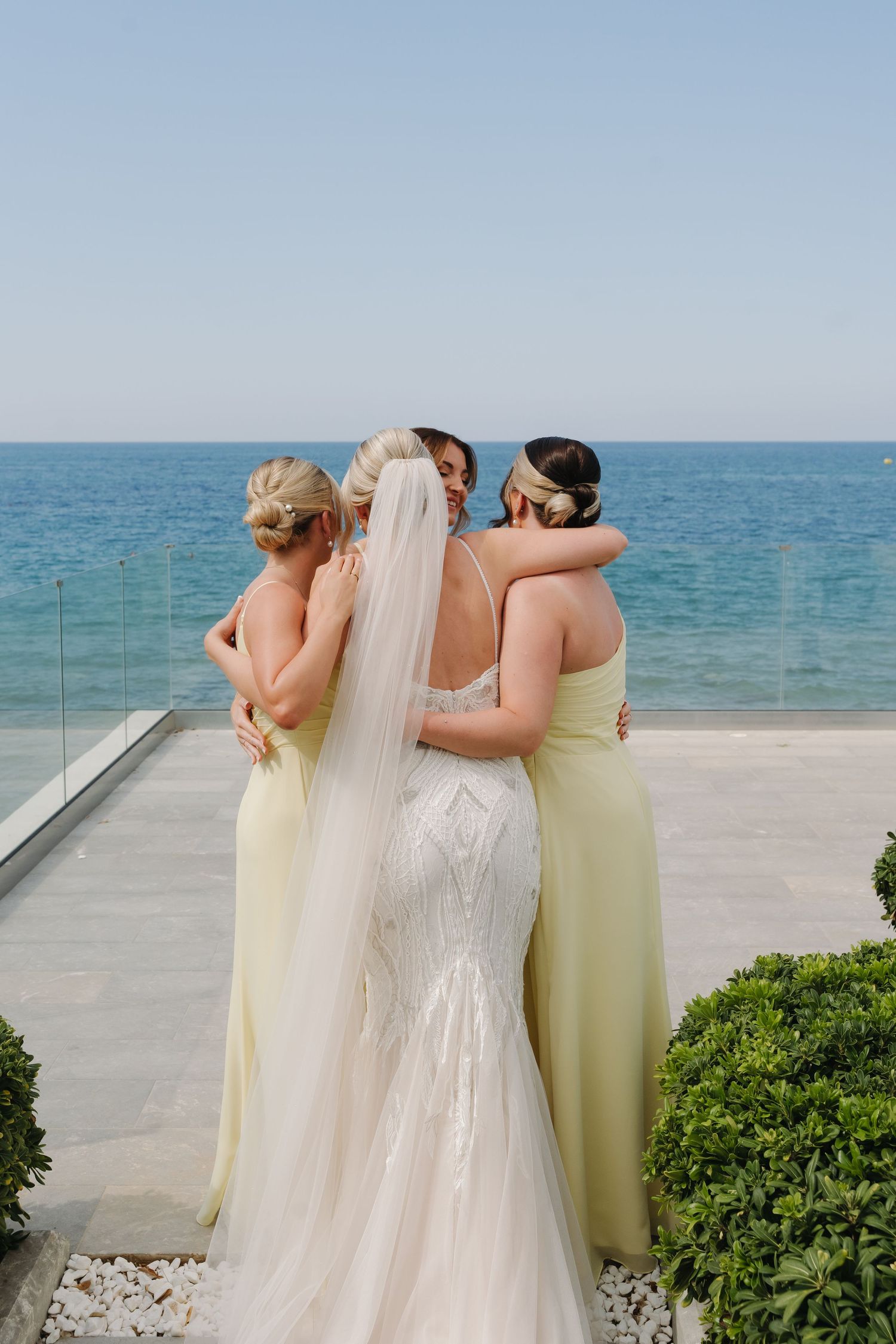 Bridesmaids in pale yellow dresses embrace around a bride in a flowing white gown at a beachside venue.
