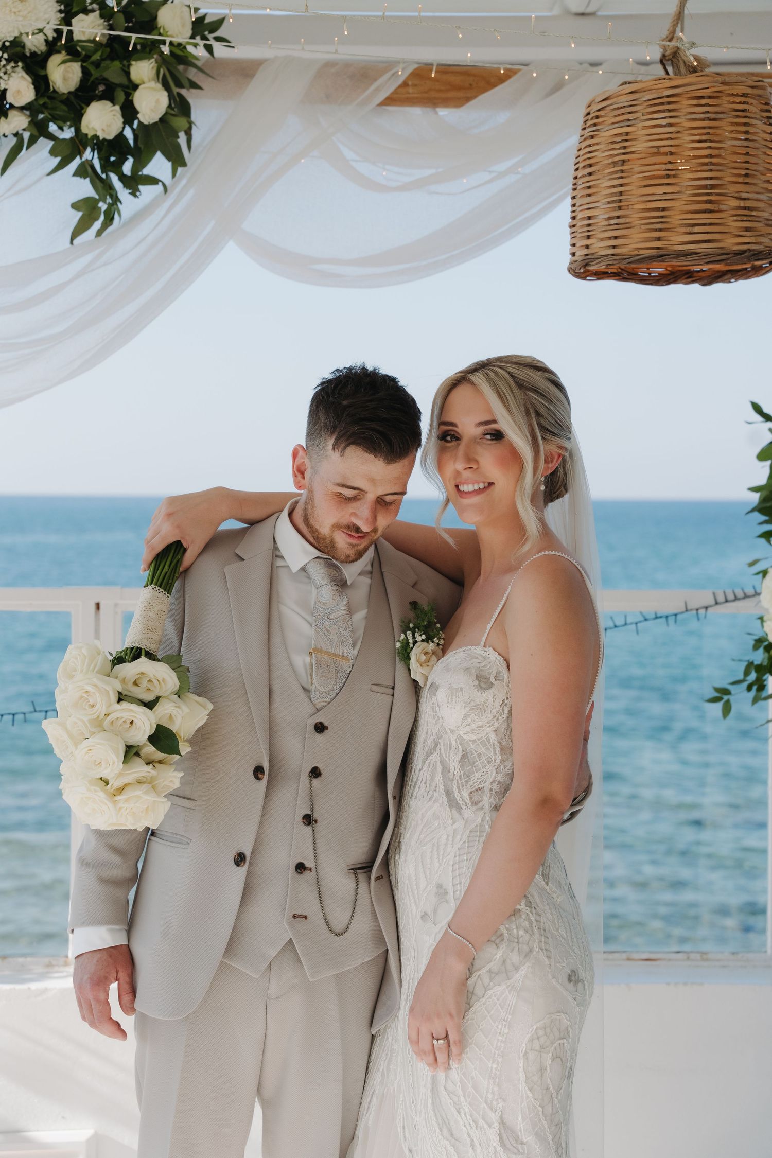 A couple shares an intimate moment at their beachfront wedding under white draping and wicker lights.