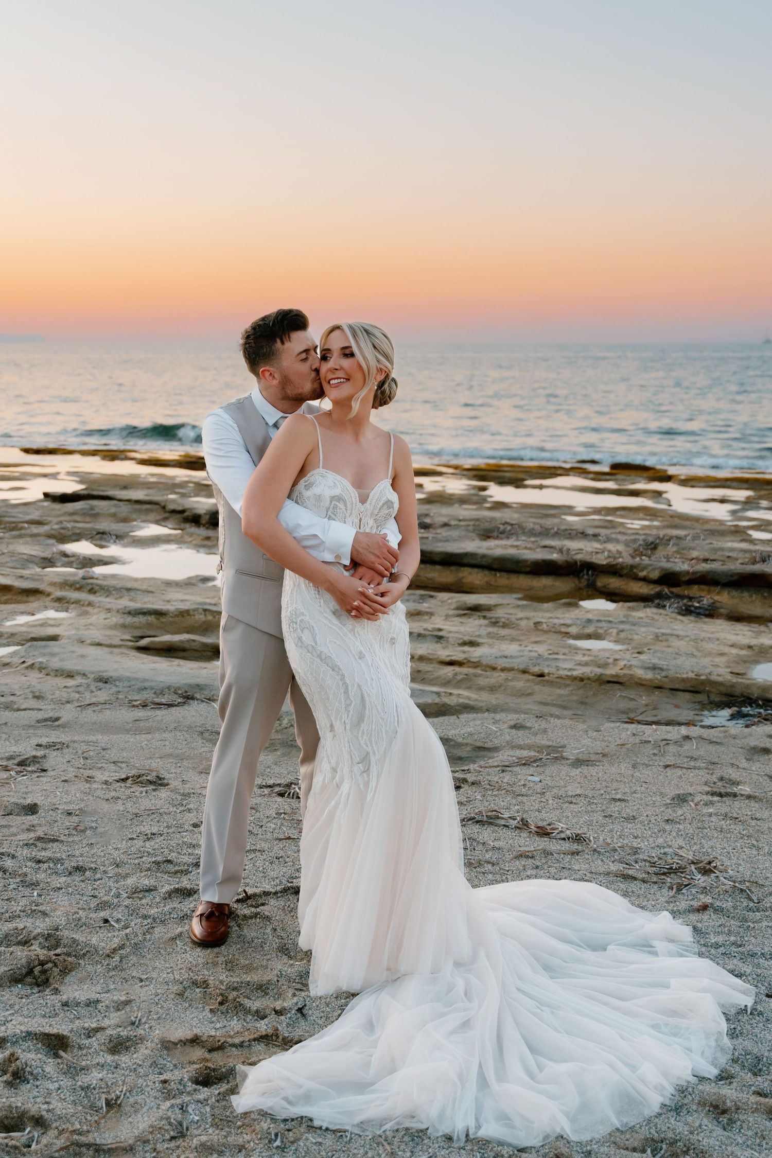 Romantic sunset beach wedding photo with flowing dress train on rocky coastline.