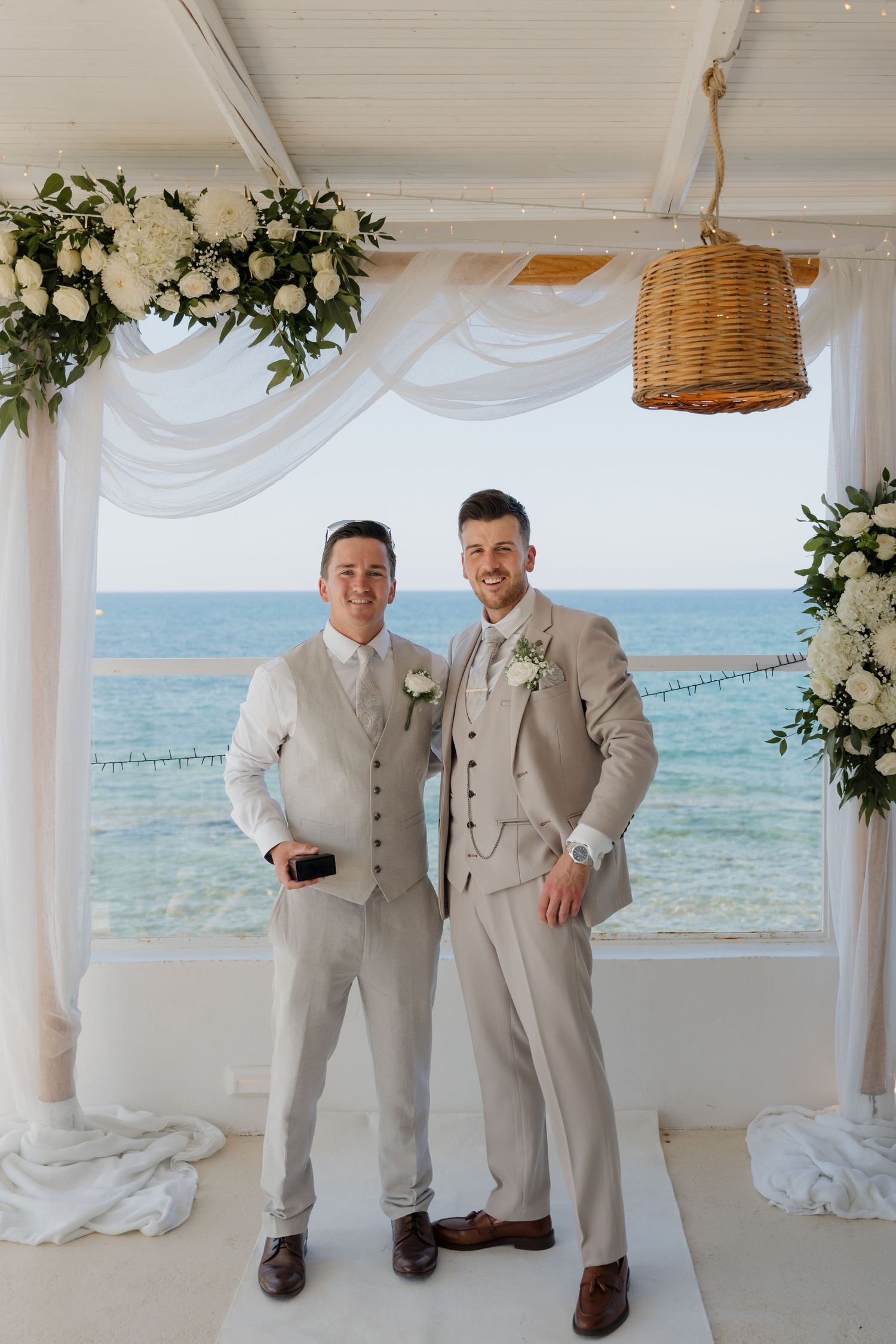 Two groomsmen in light gray suits posing against ocean backdrop with white floral arch.