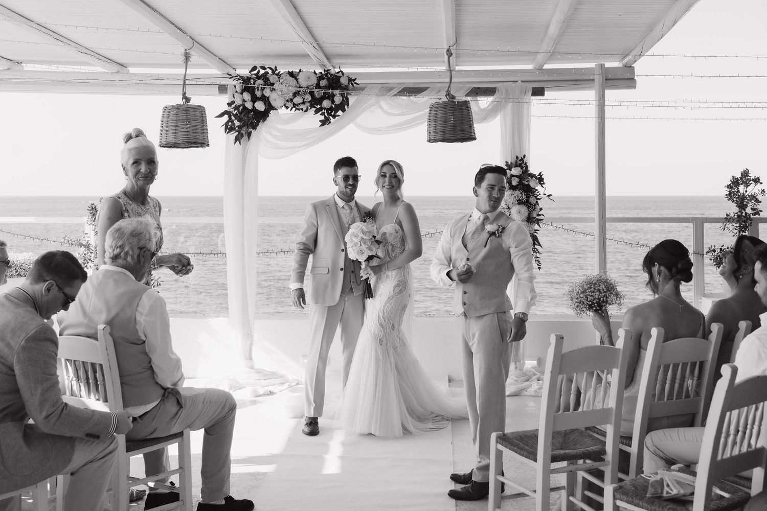 Seaside wedding ceremony under white canopy with guests seated on wooden chairs.