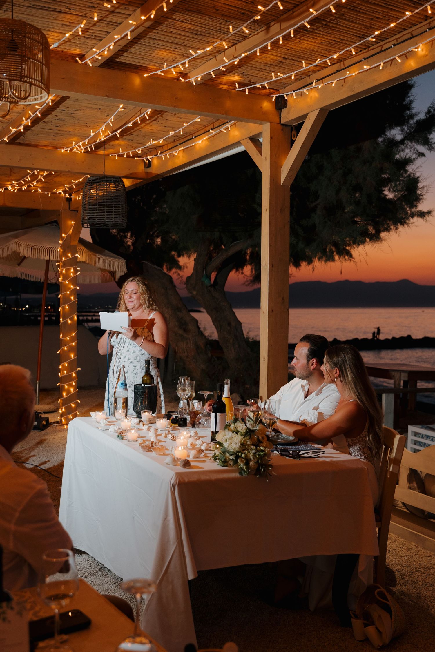 Wedding reception dinner under string lights with sunset and water view.
