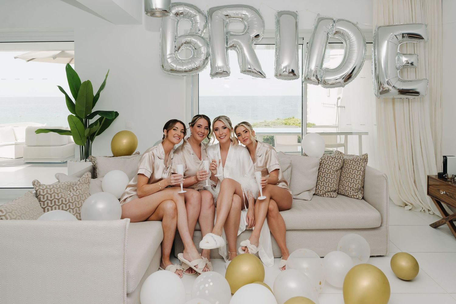 Bridesmaids sitting on white couch under silver BRIDE balloon decorations at bridal shower celebration.