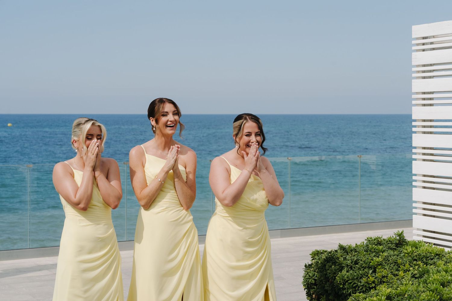 Three bridesmaids in pale yellow dresses react emotionally at beachside wedding.