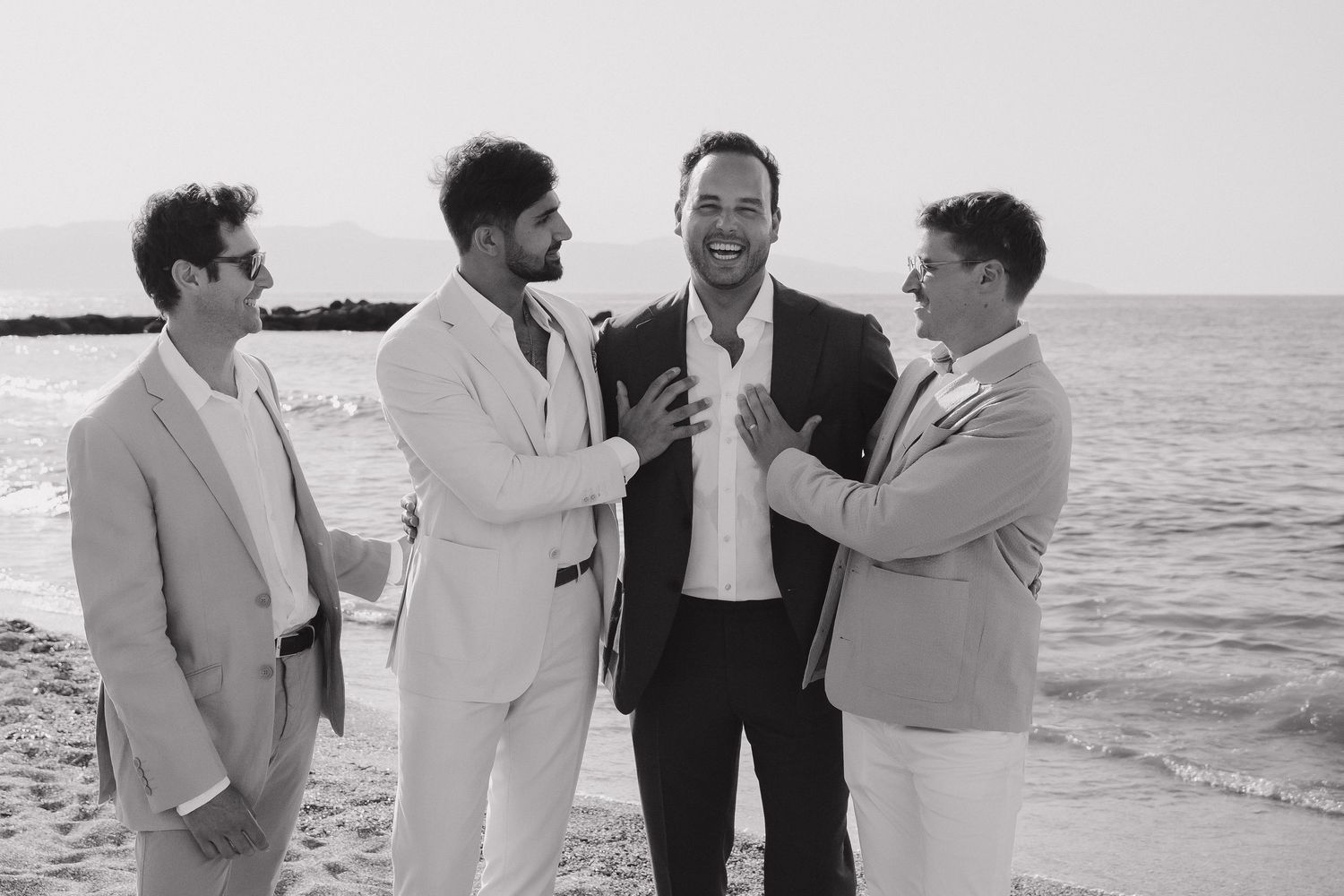 A group of men in formal attire share a candid moment together on a beach in black and white.