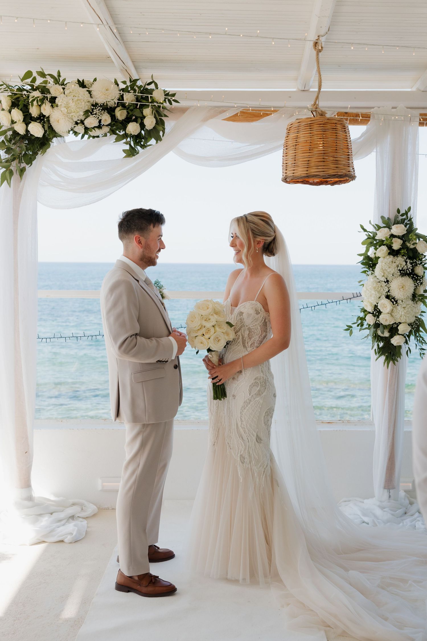 Seaside wedding ceremony with white floral arrangements and flowing curtains framing the ocean view.
