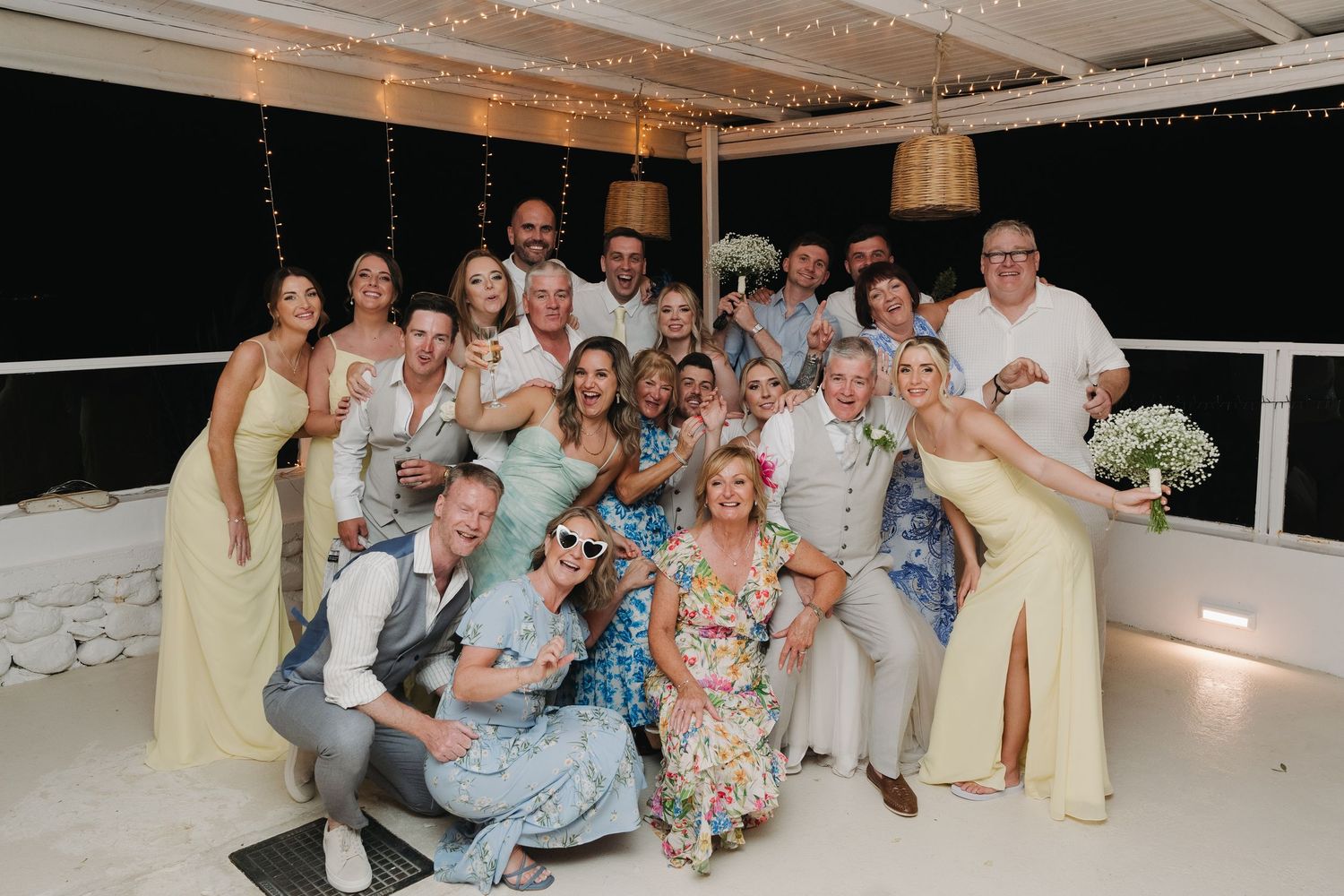 Large wedding party group photo at night on decorated oceanfront terrace.