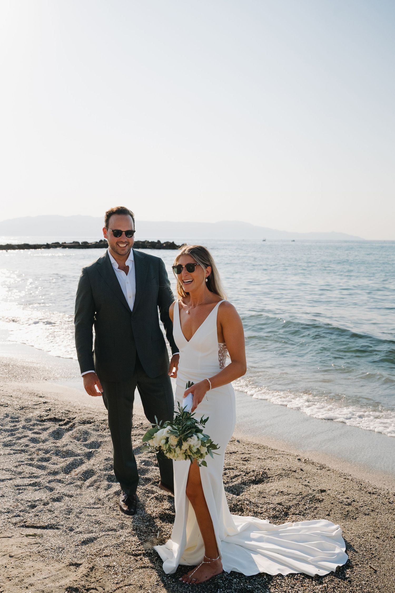Newlyweds pose together on sandy beach holding white floral bouquet.