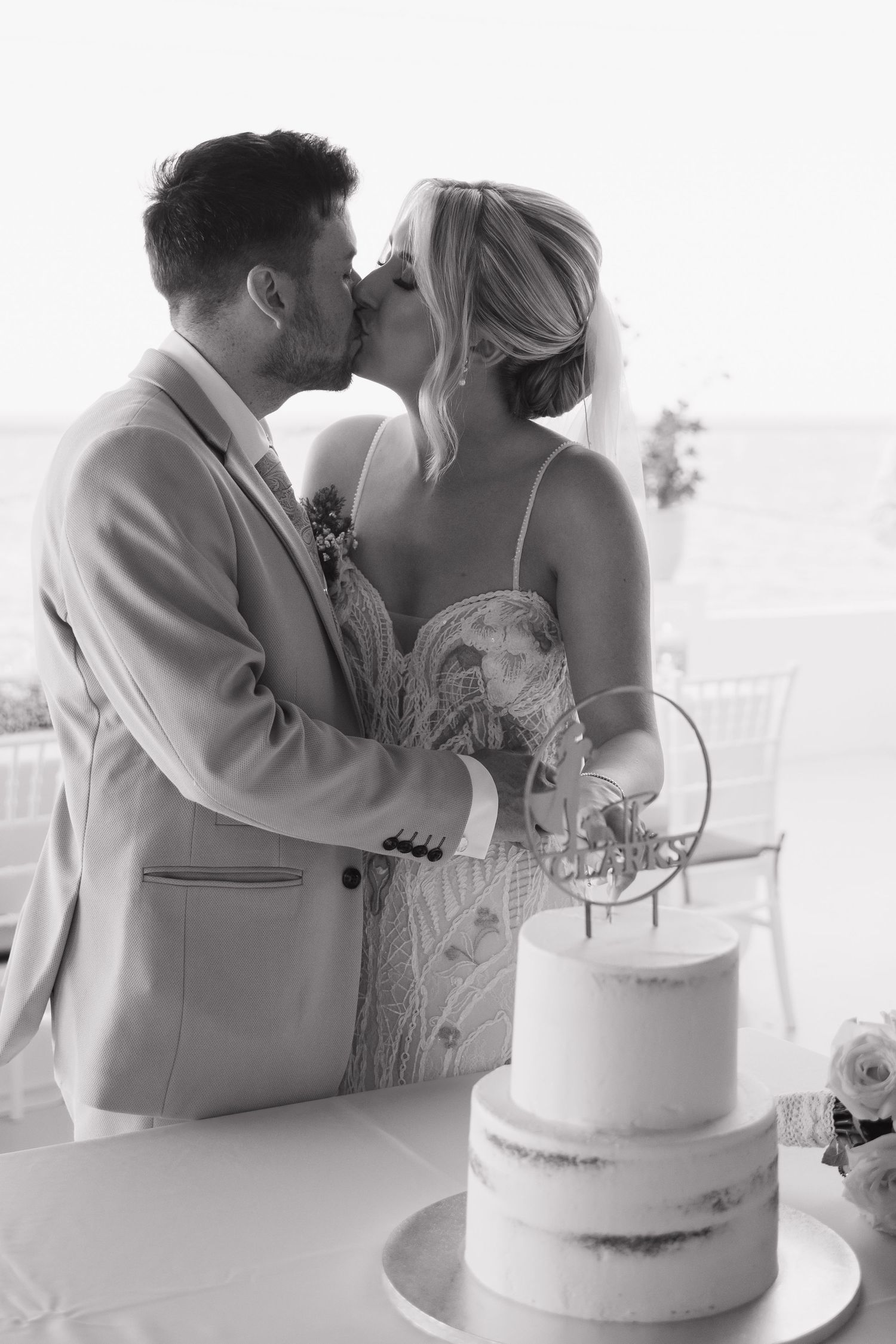 Black and white photo of newlyweds sharing a kiss beside their two-tier wedding cake.
