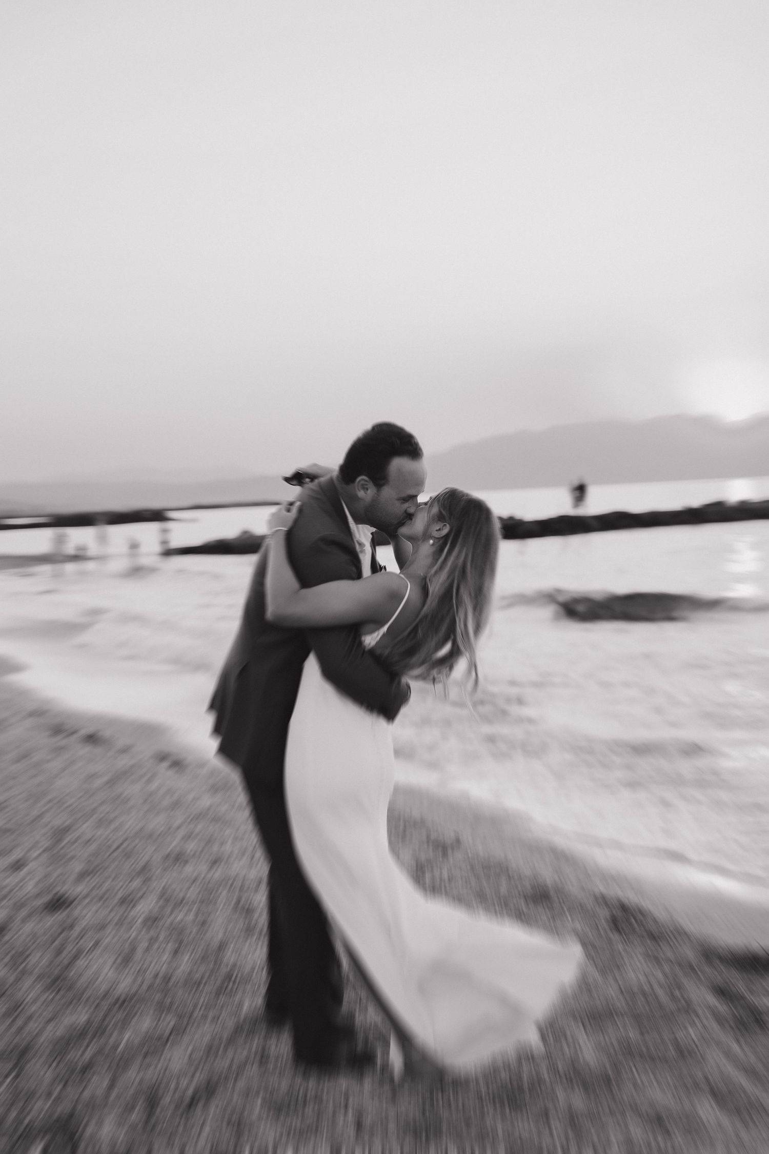 A romantic black and white beach wedding photo of a couple embracing with a flowing dress in the wind at sunset.