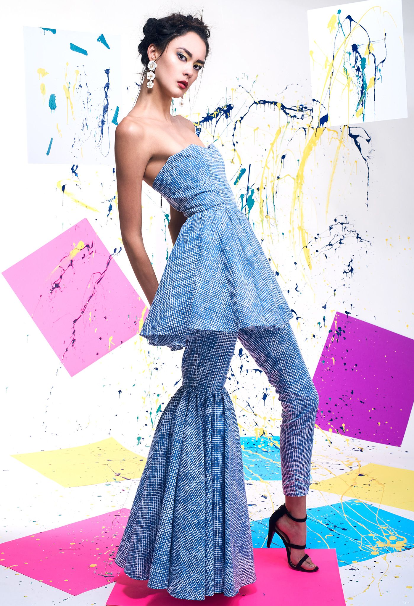 Fashion model in a blue ruffled dress poses against a colorful paint splatter backdrop.