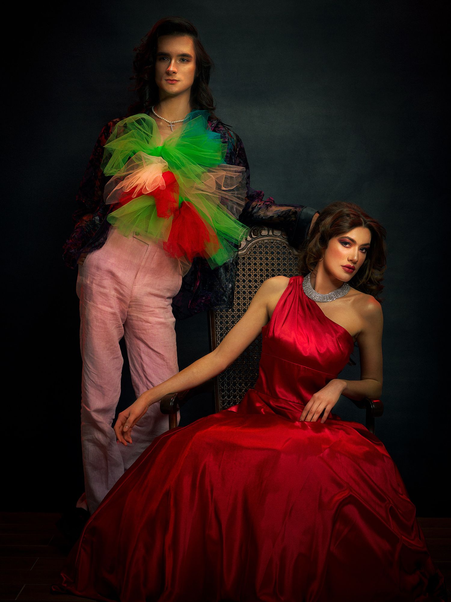 Two models pose in contrasting outfits featuring a red gown and pink pants with neon green tulle against a dark backdrop.