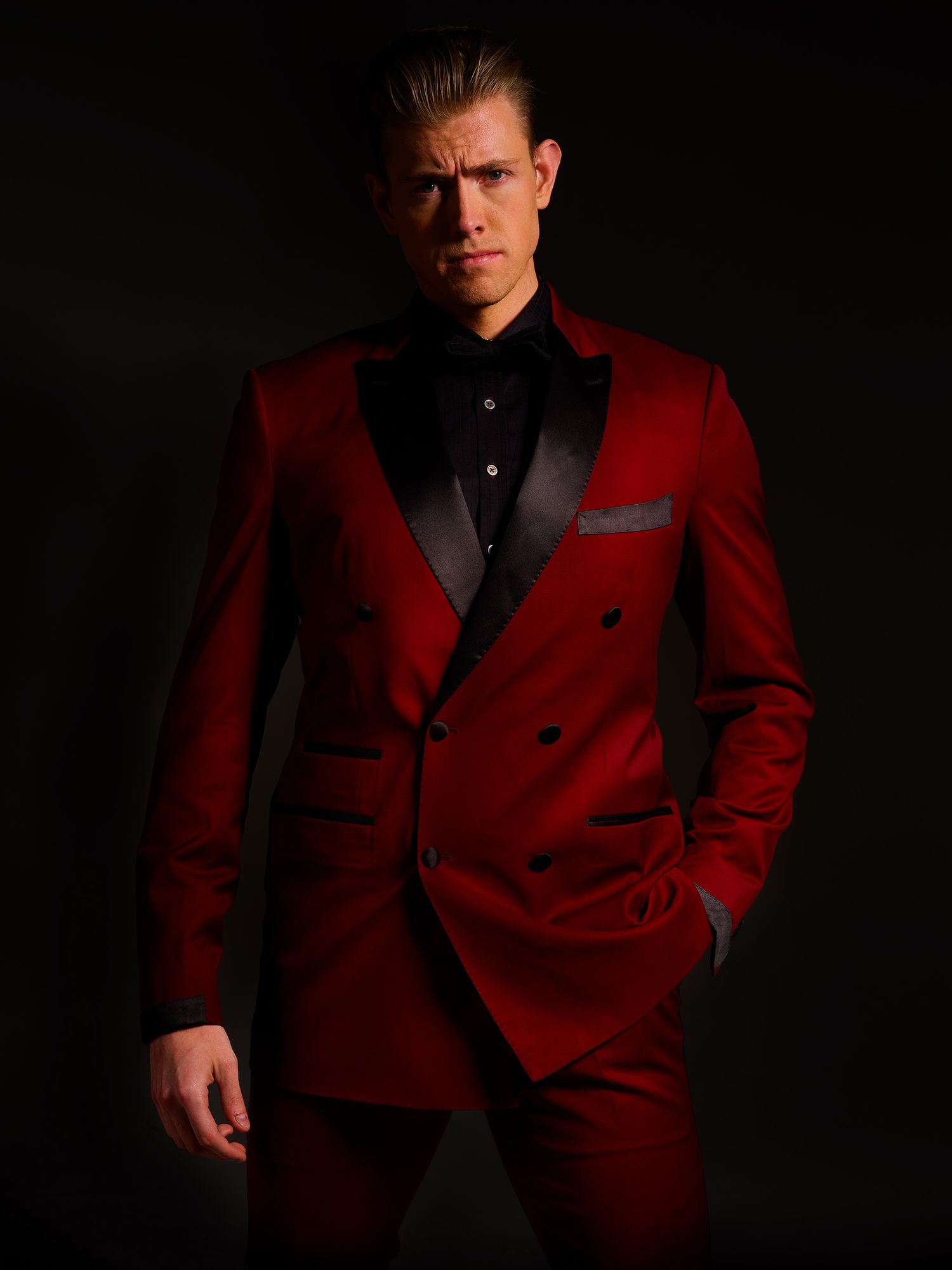 A dramatic portrait in a stylish red double-breasted tuxedo with black lapels against a dark background.