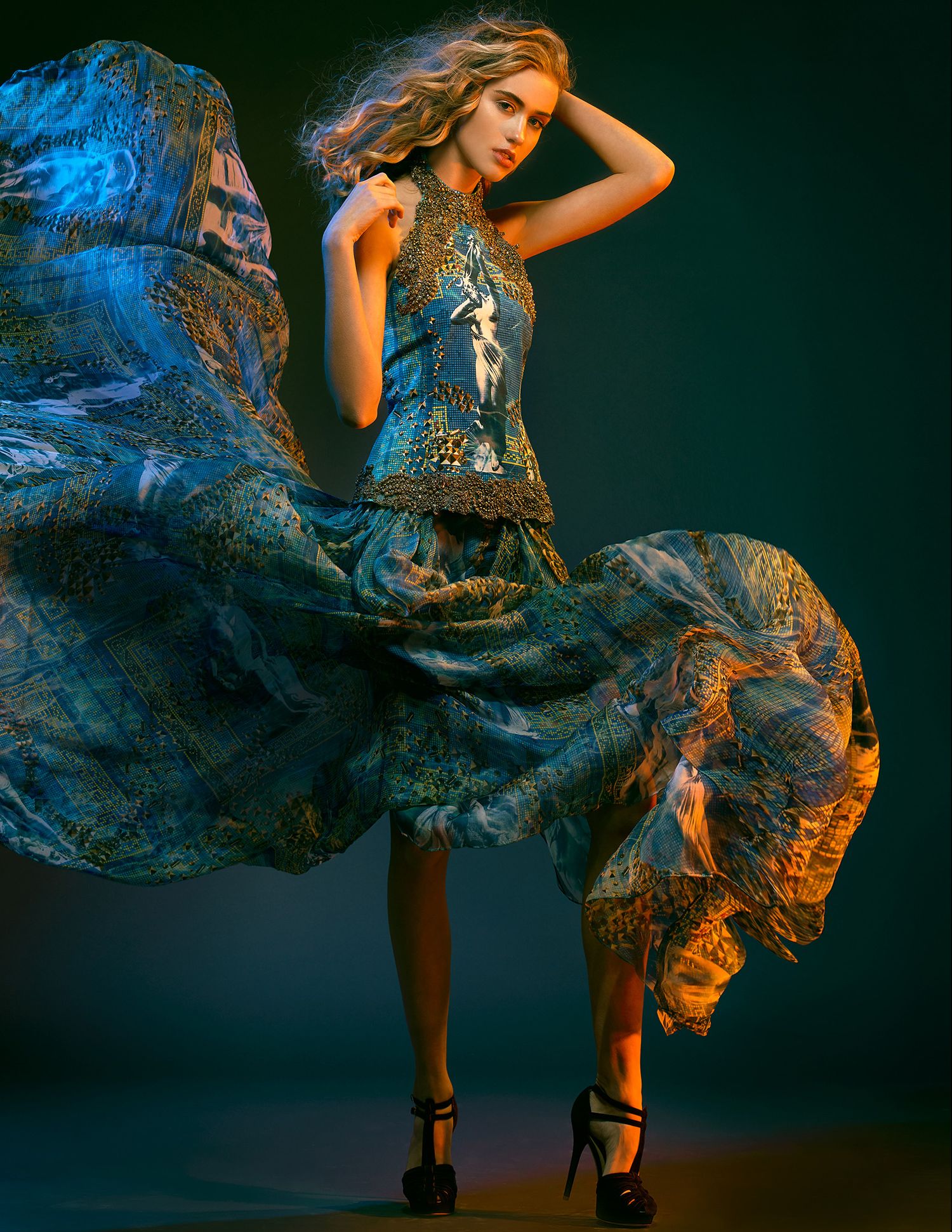 Fashion model in flowing blue patterned dress posing dramatically against dark background with artistic lighting.