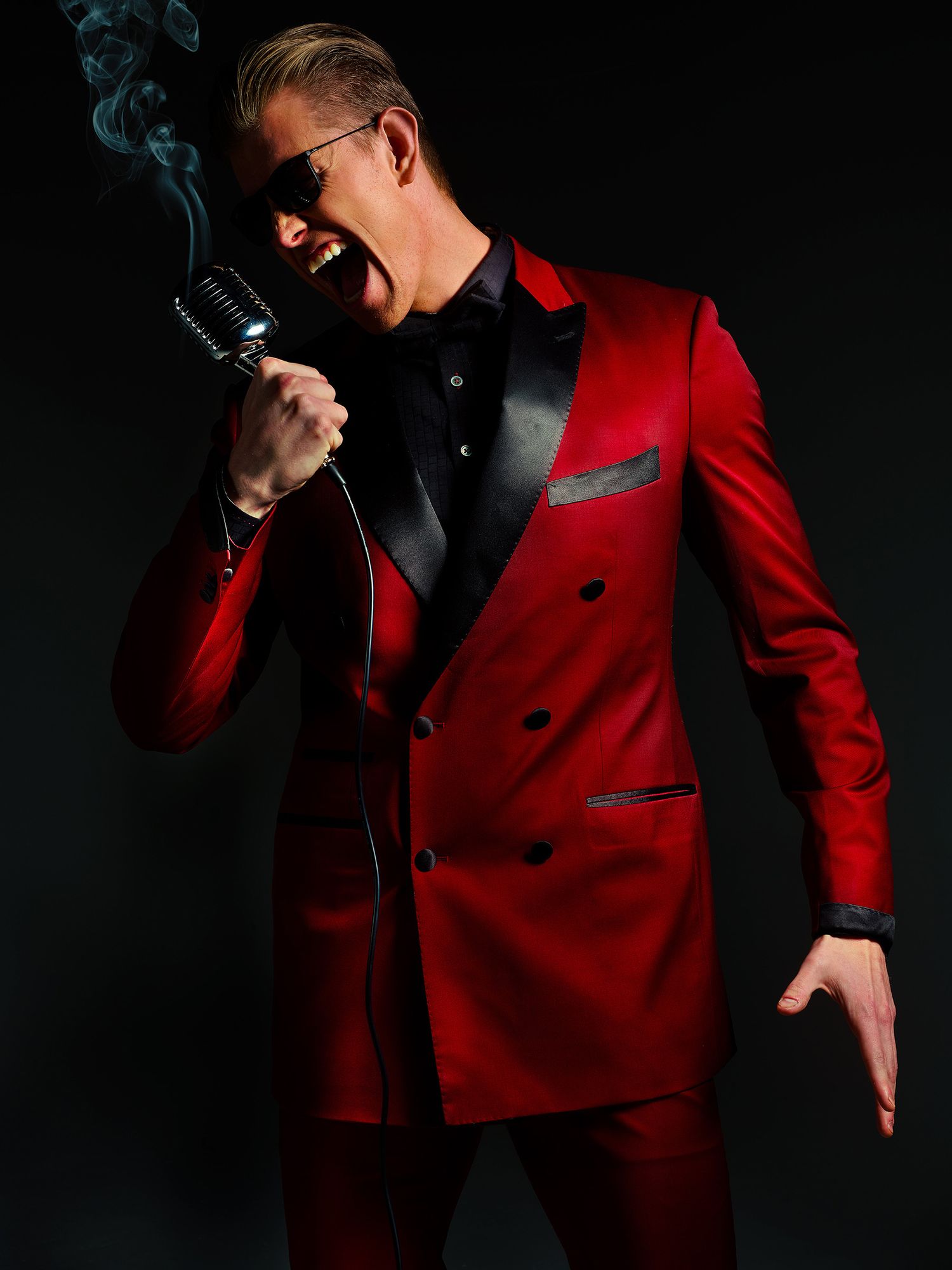 A performer in a stylish red double-breasted suit and sunglasses holds a vintage microphone against dark background.