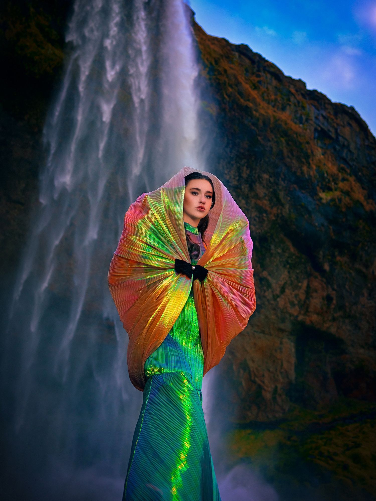 Fashion photo shows a figure in iridescent green dress with rainbow-colored fan backdrop against dramatic waterfall in Iceland.