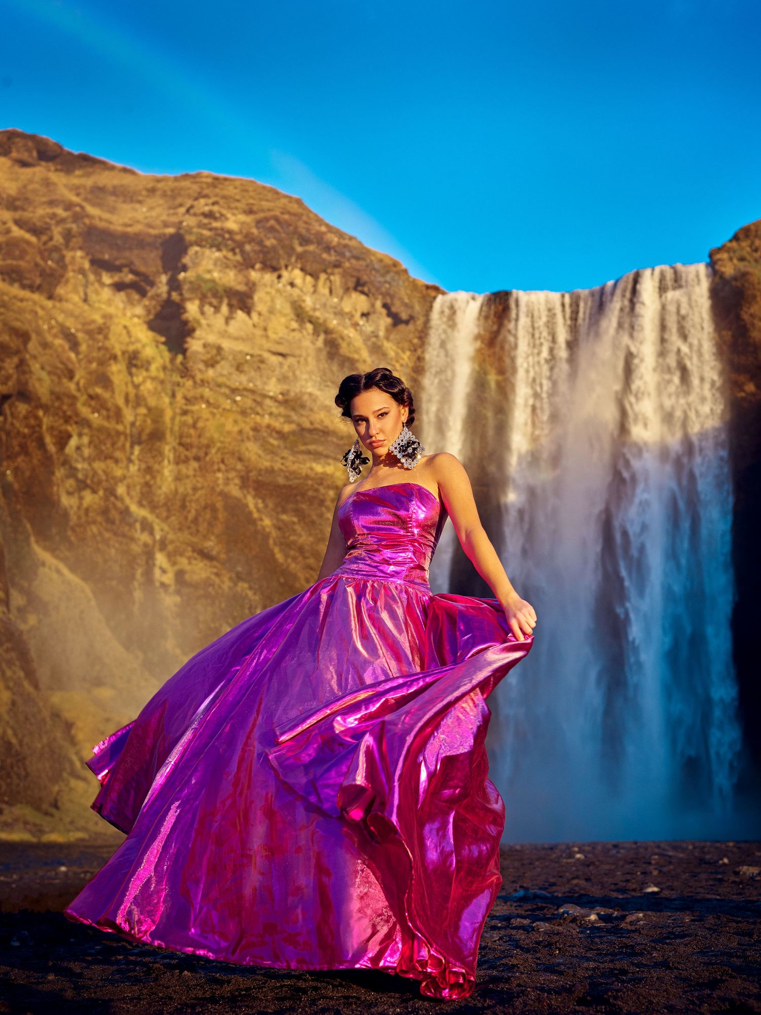 A flowing magenta ball gown catches the wind against the backdrop of a majestic waterfall and rocky cliffs in Iceland.