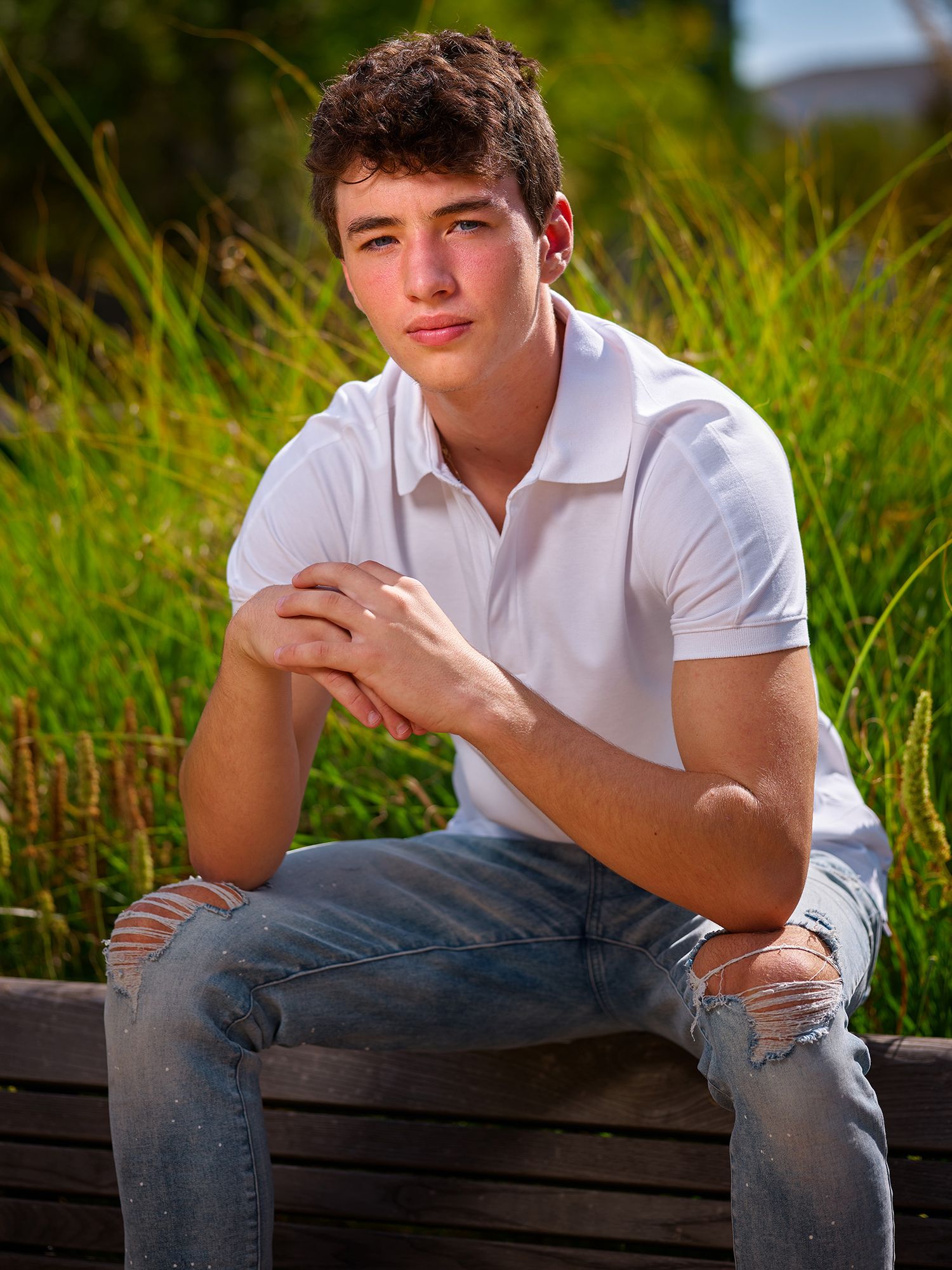 Portrait photo of someone in a white polo shirt and ripped jeans sitting outdoors among tall grass at sunset.