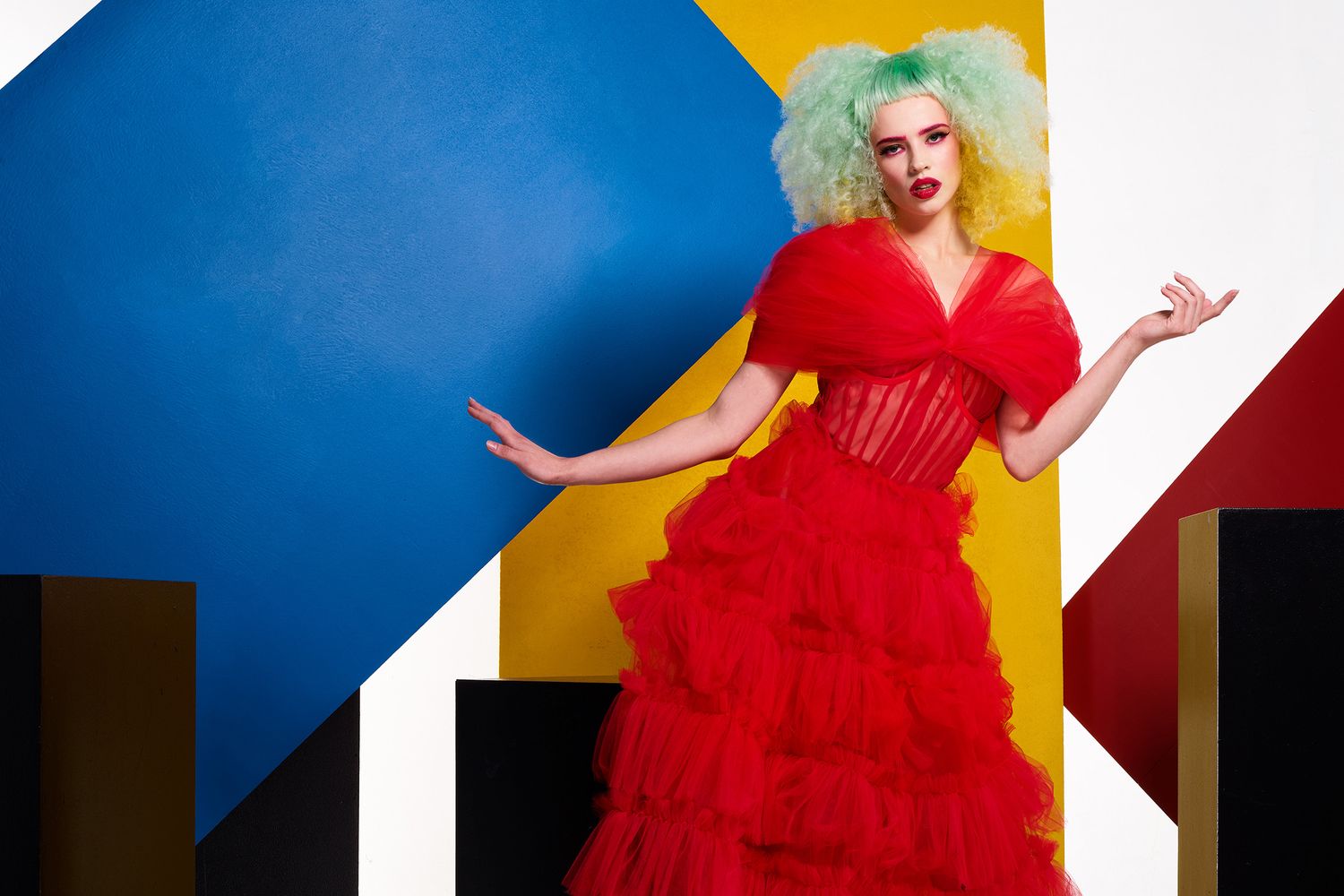 Fashion model in vibrant red dress poses dramatically against colorful geometric background.