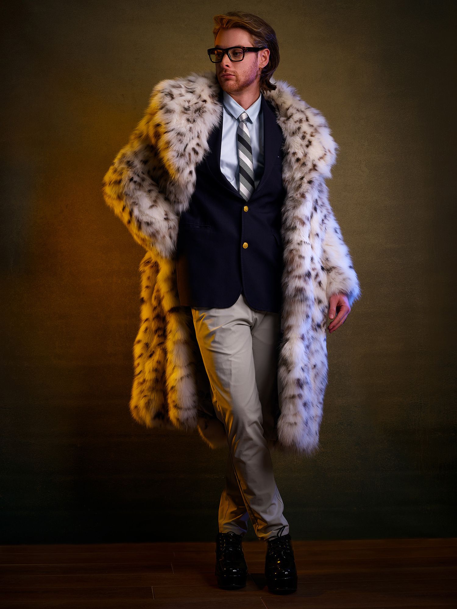 Fashion portrait showing someone in a glamorous spotted fur coat, dark blazer, khaki pants and glasses against brown backdrop.