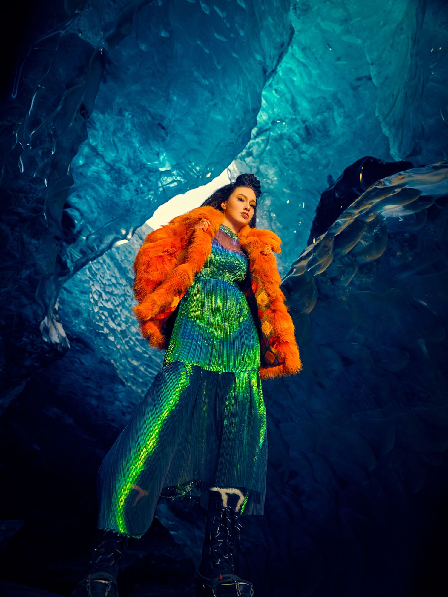 Fashion model wearing bright orange fur coat and neon green pleated dress poses against blue ice cave background.