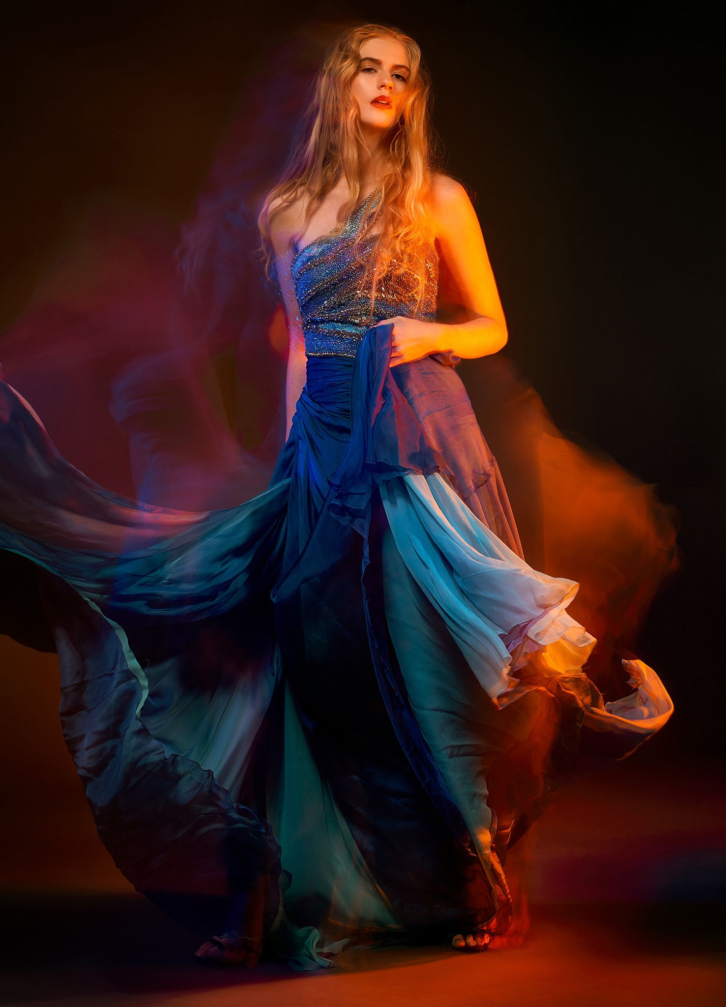 Fashion model in flowing blue evening gown dances under dramatic orange and purple lighting.