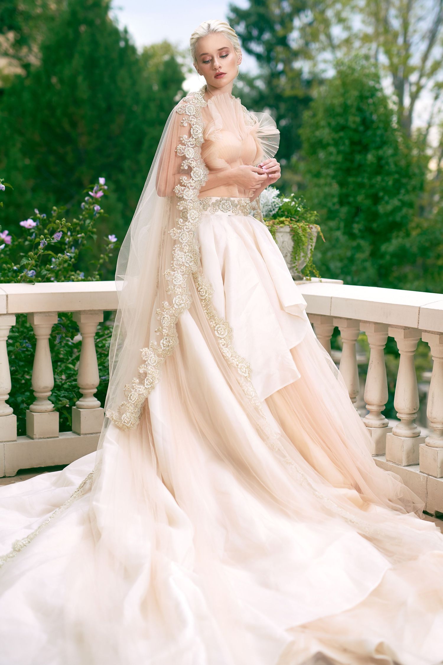 Beautiful flowy champagne wedding gown with lace details and dramatic cape billows elegantly on an outdoor balcony setting.