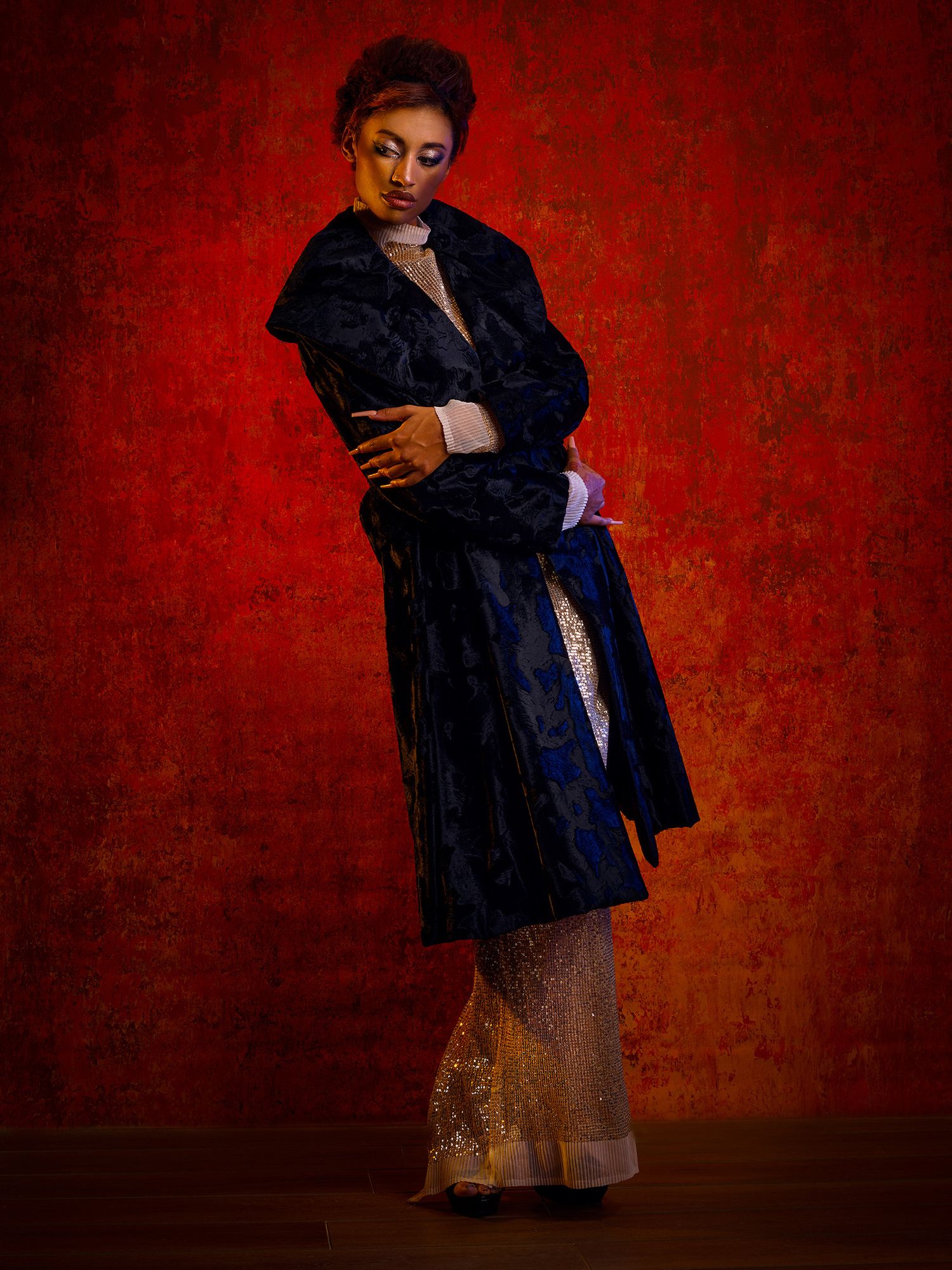 A woman wearing a dark velvet coat and gold evening dress poses elegantly against a deep red textured background.