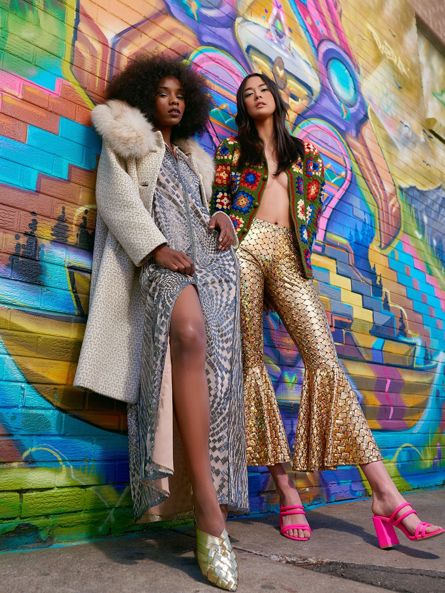 Two models pose in glamorous sequined outfits against vibrant graffiti wall wearing winter coat and bell bottom pants.